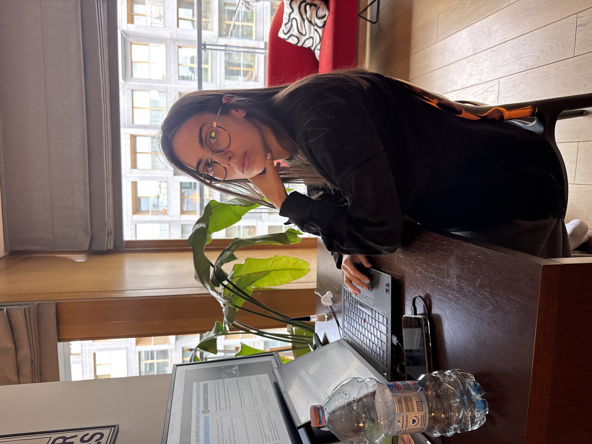 An indoor, vertically-oriented medium shot shows a young woman seated at a dark wooden desk, looking directly at the camera. She has long dark hair, wears round-framed glasses, and a dark long-sleeved top. Her right elbow rests on the desk, supporting her chin, giving her a pensive or thoughtful expression.
On the desk, to her right, is an open laptop. An external monitor stands beside the laptop, displaying text. A clear plastic water bottle with a blue cap is in the foreground, near the monitor. A dark smartphone is visible next to the laptop, appearing to be charging. Behind the setup, a large green houseplant, possibly a bird of paradise, adds a natural element.
The background features large windows, indicating a bright daytime setting, possibly late morning or early afternoon. Through the windows, a modern building with numerous rectangular windows is visible, suggesting an urban environment like Warsaw, Poland. Part of a red armchair with a white patterned cushion can be seen to the woman's left, behind her. Blinds or curtains are also visible along the windows.
The woman appears to be working or studying. The external monitor displays text in Polish, with the heading "NAJWAŻNIEJSZE ZMIANY DLA POSZCZEGÓLNYCH ZESPOŁ...", which translates to "MOST IMPORTANT CHANGES FOR INDIVIDUAL TEAMS...". The water bottle label shows "Sonnentesto" and "1L", along with what appears to be a Polish quality certification mark. A small, partially obscured sign or frame in the lower left corner displays "RS".