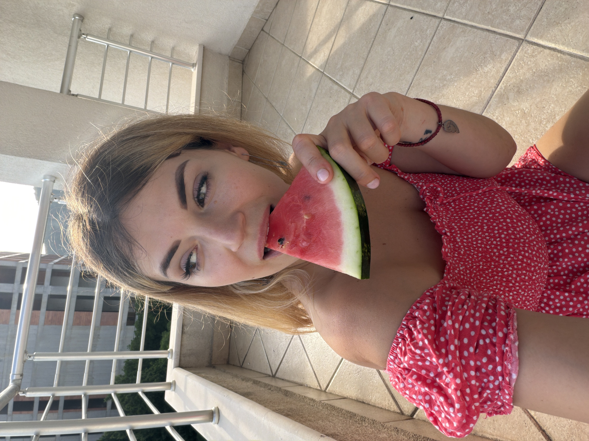 A young adult female is depicted in a high-angle, eye-level shot on an outdoor balcony in Budva, Montenegro. She has shoulder-length, light brown/blonde hair, styled down, and is looking slightly to her right with an open mouth, mid-bite into a triangular slice of watermelon. Her eyes are made up with prominent eyeliner and what appears to be light eyeshadow. A silver chain-link earring is visible on her left ear. She wears a red, off-the-shoulder dress with white polka dots, featuring a shirred bodice and puffed sleeves. Her right hand holds the watermelon slice, revealing light pink nail polish. A dark red braided bracelet with a silver half-heart charm is on her left wrist. The watermelon displays vibrant red flesh, black seeds, a thin white rind, and green outer skin. The balcony floor is paved with light beige square tiles. A polished metal railing with horizontal bars runs along the balcony's edge. In the background, under clear skies and bright daylight, multiple multi-story concrete buildings in various stages of construction or partial completion are visible. Green trees and foliage are situated below and between the structures.
