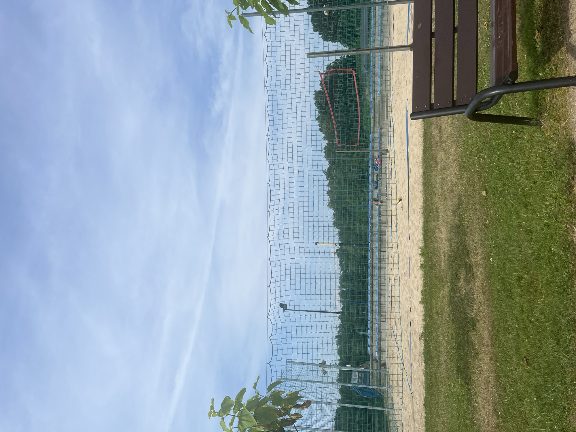 This media file depicts a sandy court with a net, likely for a sport like volleyball or beach tennis, set against a backdrop of trees and a bright blue sky with scattered clouds. The scene is oriented vertically, with the sky occupying the left side and the court and its surroundings on the right. A dark wooden bench with black metal legs is visible in the foreground on the right, suggesting a place for spectators or players to rest. The court itself is sandy, with a net stretched across it. Behind the net, rows of blue seats are visible, indicating a designated spectator area. A red rectangular frame is placed on the court in the distance, possibly marking a practice area or a specific game element. The overall impression is of an outdoor recreational facility. The lighting suggests it is daytime, with the sun casting shadows, and the weather appears clear and pleasant. There are no visible people engaged in any activity, but the presence of sports equipment and seating implies the potential for games or practice sessions. The environment seems to be a park or a sports complex in a natural setting, given the abundance of trees. No text is visible in the image.