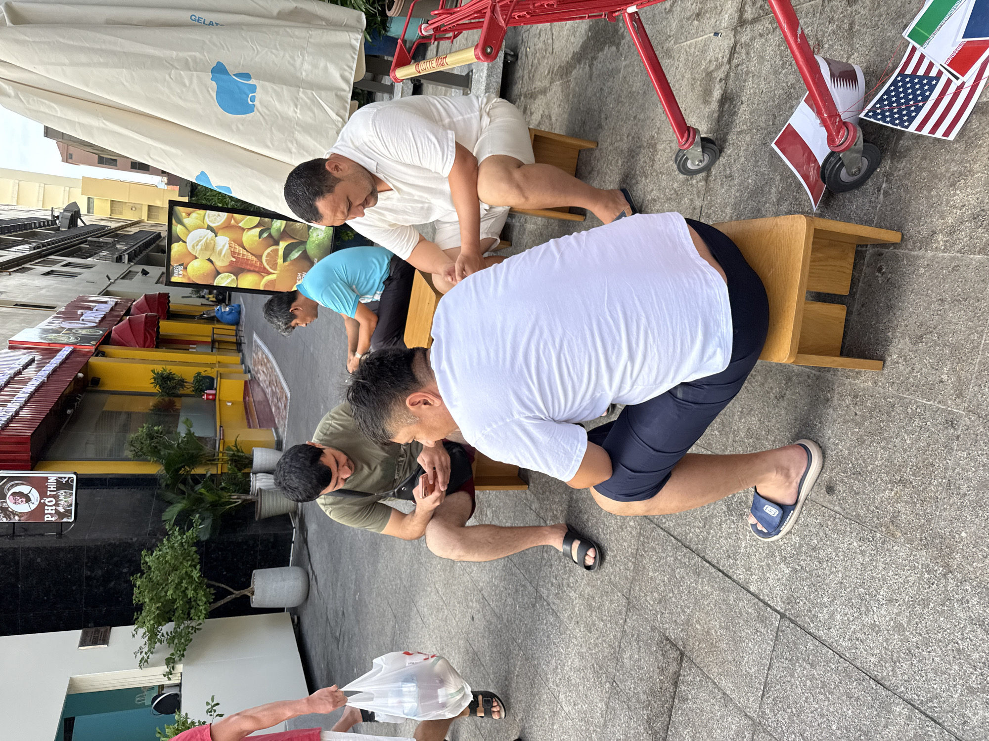 In Nha Trang Ward, Vietnam, four men are gathered on a paved walkway. Two men, dressed in casual attire, are seated on wooden stools, one looking at his phone and the other conversing with the man beside him. A third man, wearing a light-colored shirt and shorts, sits on a stool in the foreground, facing away from the camera. The fourth man, also in casual clothing, is seated on the ground in the background. A shopping cart with red accents and a large, cream-colored umbrella are visible to the right, partially obscuring a building with a yellow facade and awnings. Several potted plants are placed along the walkway, and signage for "PHO THIN HANOI" is visible on a building. The scene appears to be a public outdoor area in a commercial district.