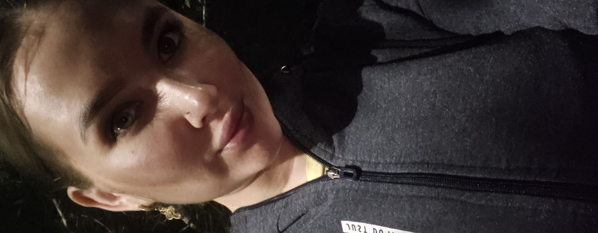 A close-up, low-angle selfie shows a woman from the chest up, outdoors at night. She has fair skin, dark hair pulled back, and dark makeup around her eyes. She is wearing a dark gray zippered hoodie over a yellow shirt and a small, gold-colored flower-shaped earring in her left ear. Her right ear is not visible. The background is dark and out of focus, with some foliage visible on the left. The woman's facial expression is neutral, with her lips slightly pursed. The image appears to have been taken in the city of Semey, Kazakhstan, although no specific landmarks are visible.