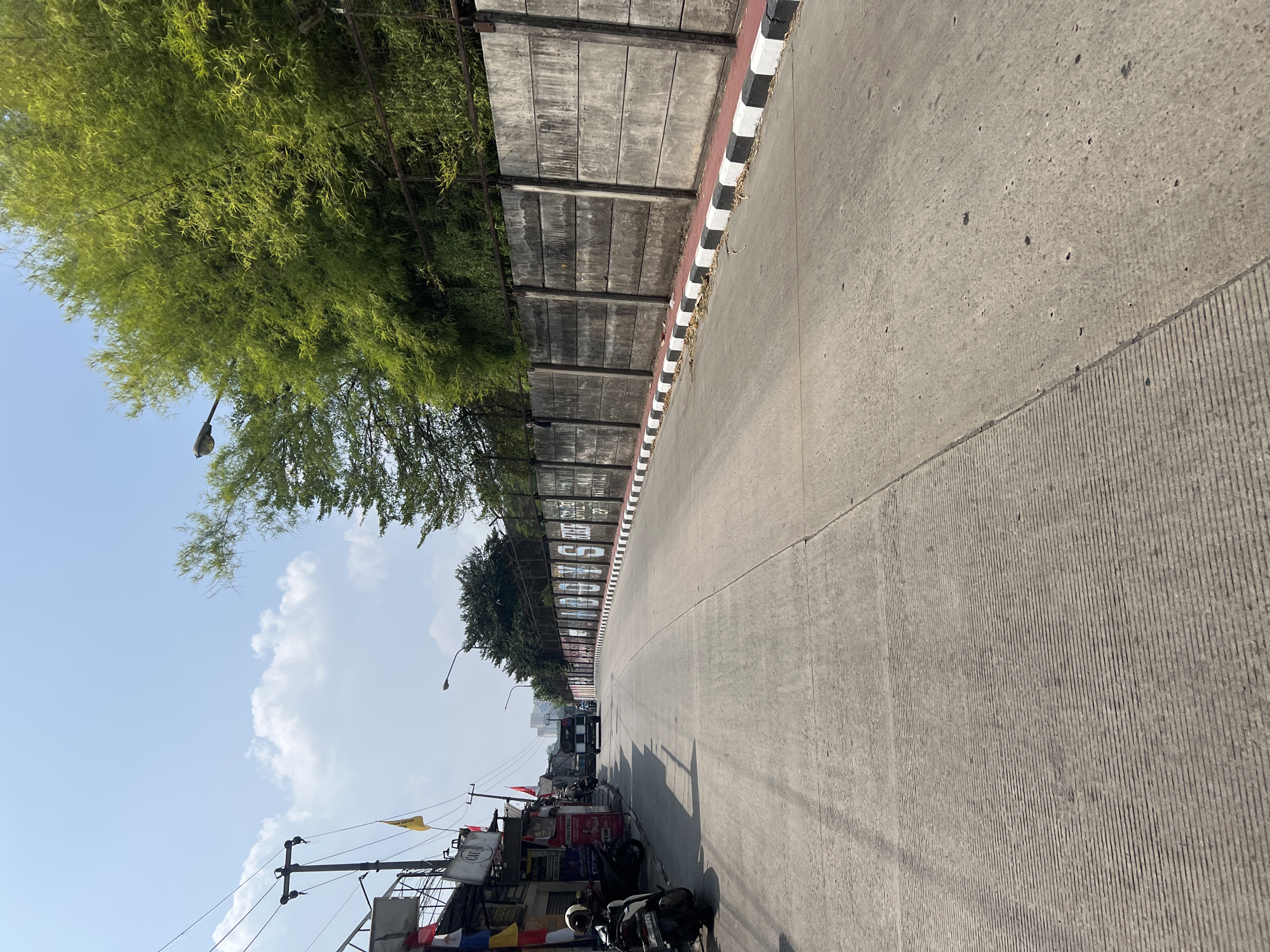 The image depicts a street scene in Gunung Putri, Indonesia, on a sunny day. A concrete road with textured lanes runs through the center of the frame, leading towards a distant urban landscape. On the left side of the road, a row of buildings with colorful flags and awnings is visible, with several parked motorcycles. A utility pole with wires stands near the buildings. On the right side, a tall, weathered concrete wall with vertical supports is lined by a red curb and a black and white striped border. Lush green trees, including bamboo, grow behind the wall. A street lamp is positioned above the wall, and another can be seen further down the road. A truck is driving away from the camera in the middle of the road, and a few other vehicles and individuals are present in the distance. The sky is blue with scattered white clouds.