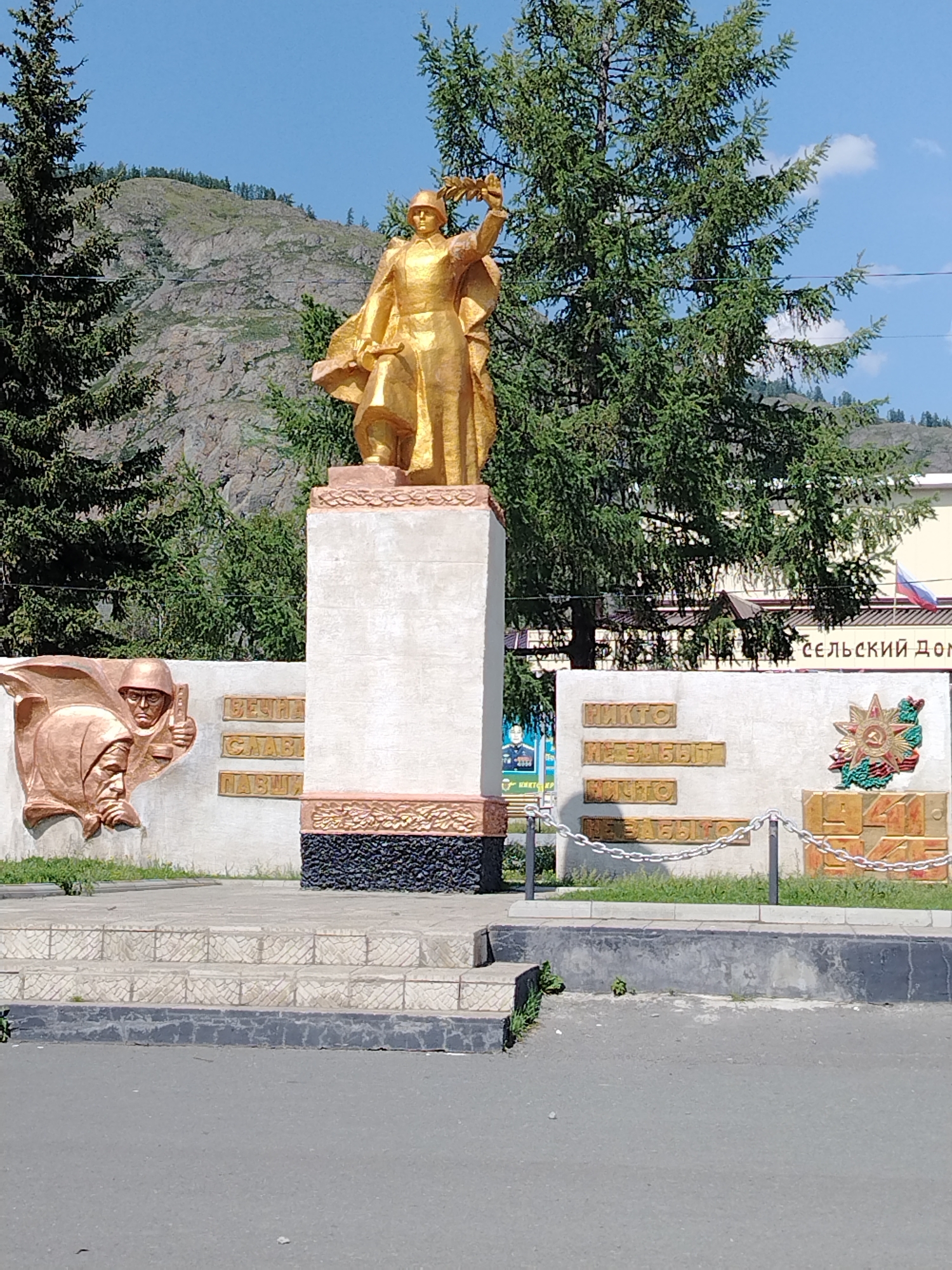 A golden statue of a soldier holding a wreath stands prominently in front of a concrete monument. The monument has various inscriptions and relief sculptures. To the left of the statue, there are relief sculptures of two figures, one wearing a helmet and the other a headscarf. Inscriptions in Russian are visible on the monument's wall. To the right of the statue, there is a Russian flag visible on a building in the background, which has Cyrillic text that reads "СЕЛЬСКИЙ ДОМ" (Rural House). The statue and monument are set against a backdrop of a rocky mountain, green trees, and a blue sky with some clouds. The scene is observed from a street level, with a paved road in the foreground and steps leading up to the monument. The overall setting appears to be a public space, likely a memorial or monument site in the city of Aktash, Russia.