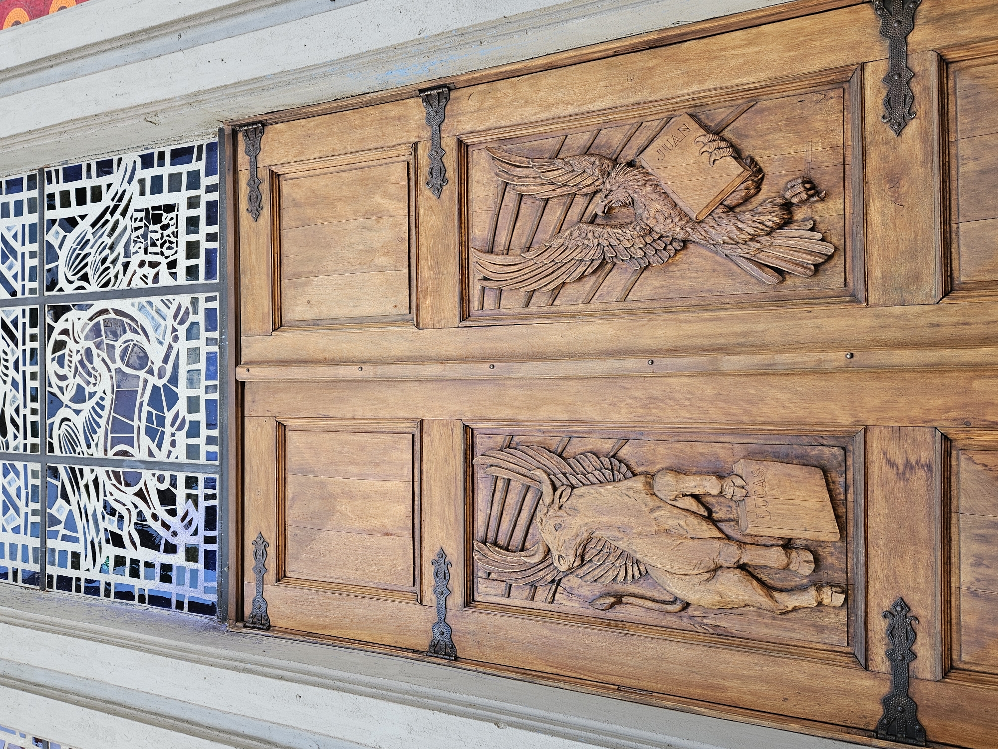 This image presents a detailed view of an architectural feature, likely part of a building entrance or facade, combining carved wood and stained glass. The scene focuses on a section of a wooden door adjacent to a vibrant window panel.
The wooden door features two prominent carved panels, one above the other, each depicting a symbolic figure holding a book. The upper carving portrays a majestic eagle with outstretched wings, grasping an open book in its talons. The name "JUAN" is clearly carved on the pages of this book. Below, the second carved panel illustrates a winged ox or bull, also holding an open book, with the name "LUCAS" inscribed on its pages. These figures are traditional symbols of the Evangelists John and Luke, suggesting the building is of religious significance, such as a church. The wood has a warm, medium-brown finish, highlighting its natural grain and the depth of the carvings. Decorative, dark metal fittings or hinges are visible between the wooden panels.
To the left of the wooden door, a striking stained-glass window is installed. It is composed of numerous small, irregular pieces of blue, white, and clear glass, forming intricate, abstract patterns that might suggest stylized figures or motifs. The entire structure is framed by a light-colored, perhaps stone or concrete, architectural molding. The image is brightly lit, likely by natural daylight, showcasing the fine details of both the carvings and the stained glass. No activity is occurring, but the craftsmanship of the elements is a central focus.