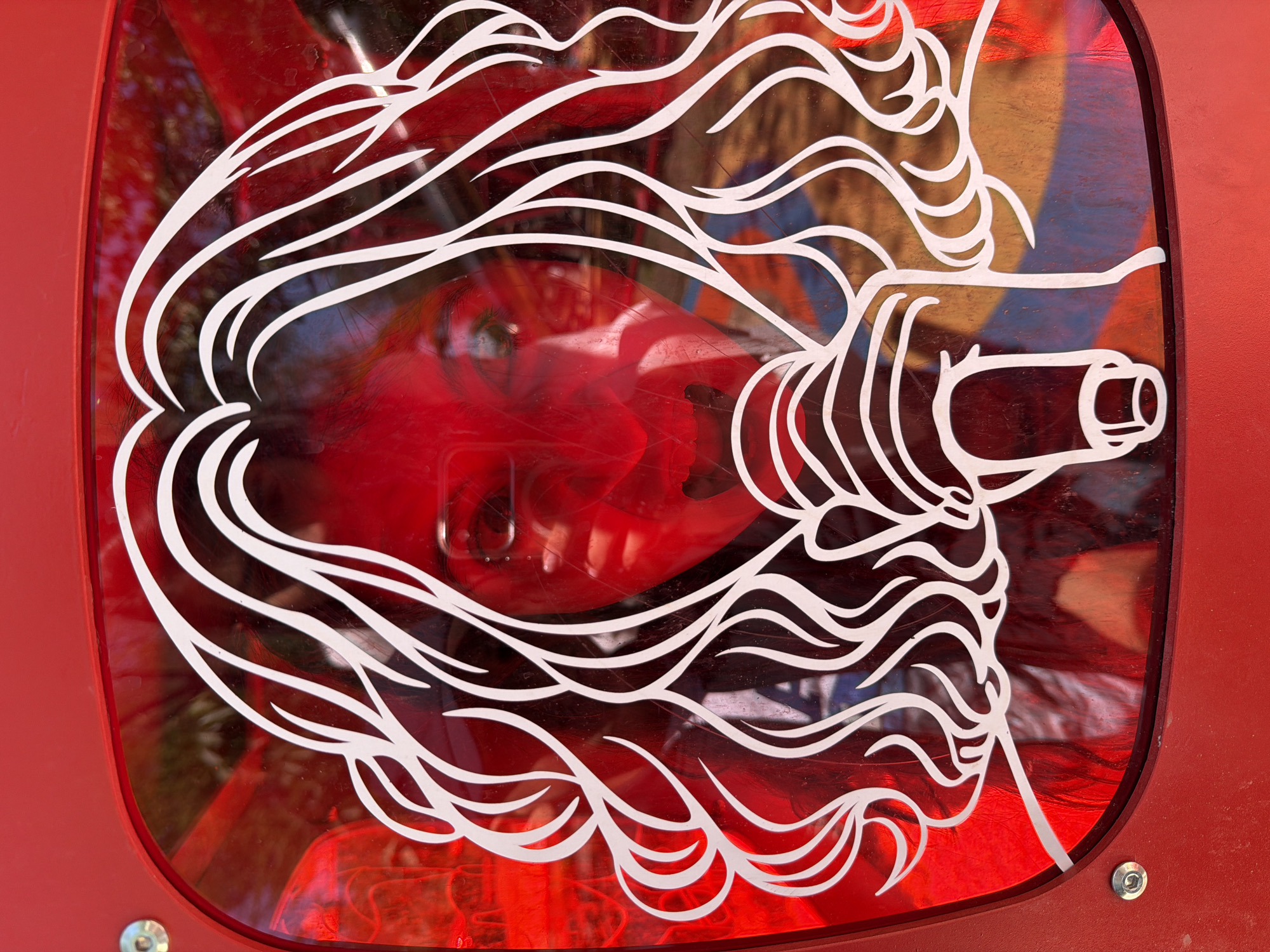 A close-up shot features a red rectangular structure containing a semi-transparent window. Inside the window is a red object, possibly a face or sculpture, obscured by a white, swirling, graphic overlay reminiscent of hair or flowing lines. This overlay creates a dynamic visual effect, partially concealing the object behind it.
The setting is Yerevan, Armenia.