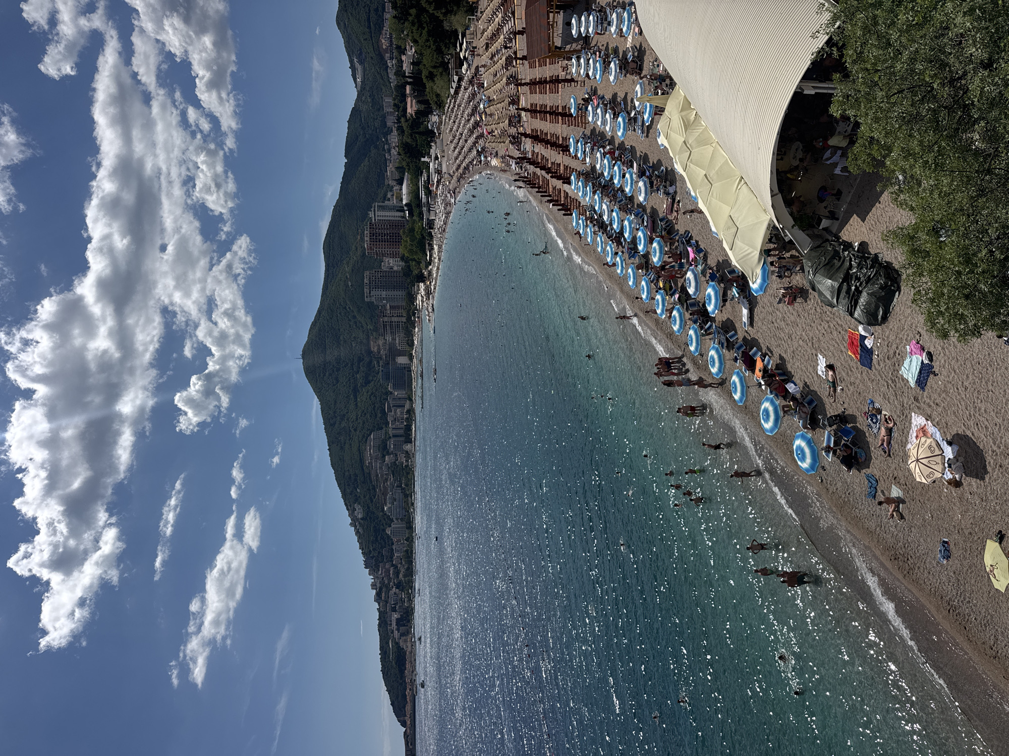 This is a high-angle, full-shot photograph of a crowded beach on a sunny day, likely in Budva, Montenegro. The scene captures a wide expanse of the Adriatic Sea with its surface shimmering under the sunlight. Along the coastline, a densely packed beach is visible, lined with rows of lounge chairs and umbrellas. Numerous people are present on the beach, some sunbathing on towels, while others are scattered among the chairs or walking along the shore. A significant number of individuals are also in the water, swimming and wading.
In the background, a hillside covered in lush green vegetation rises steeply, dotted with numerous buildings, forming a town or city. The buildings appear to be a mix of residential and commercial structures, with some under construction. The sky above is a vibrant blue, partially obscured by scattered white cumulus clouds, with sunbeams breaking through.
The foreground features a section of the beach with more lounge chairs and umbrellas, and a portion of a beachside establishment with a curved, light-colored roof. Several trees with green foliage are visible in the lower right corner, framing the scene. The overall impression is one of a popular tourist destination during peak season.