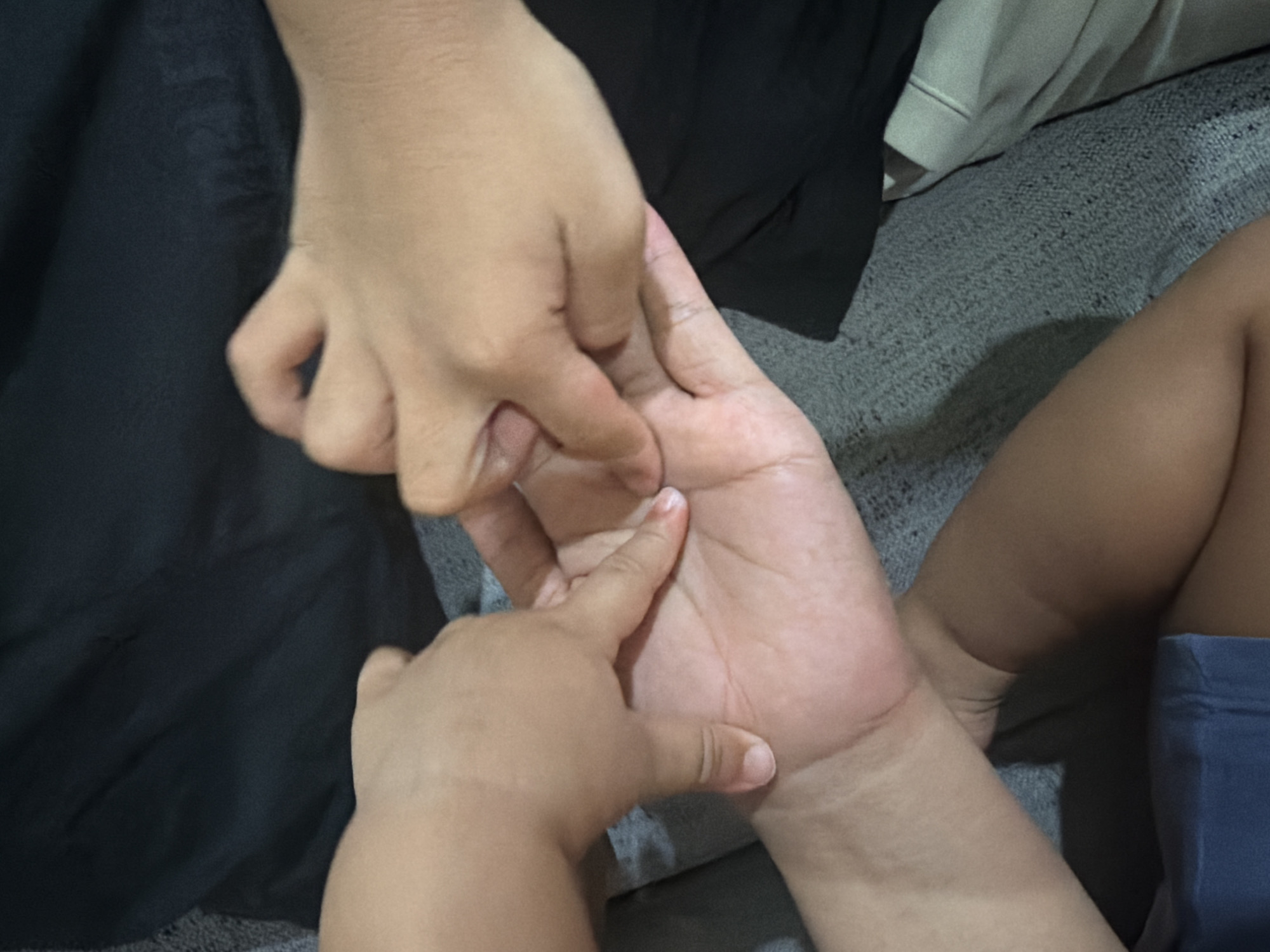 The image shows a close-up view of two people's hands interacting. One hand, appearing to be that of a child, is gently touching or holding the palm of another, presumably an adult's hand. The larger hand is open, displaying its palm and fingers. The smaller hand is positioned over the larger palm, with one finger lightly pressing down. The background is blurred, but suggests a domestic setting with fabric textures, possibly a sofa. One person is wearing dark clothing, and another is wearing blue shorts. The lighting is dim, suggesting it could be indoors during the evening or in a dimly lit room. There is no visible text in the image. The interaction appears to be tender and affectionate, possibly a parent comforting a child or a sibling's gentle touch. The focus is entirely on the hands, highlighting the connection between the individuals.
