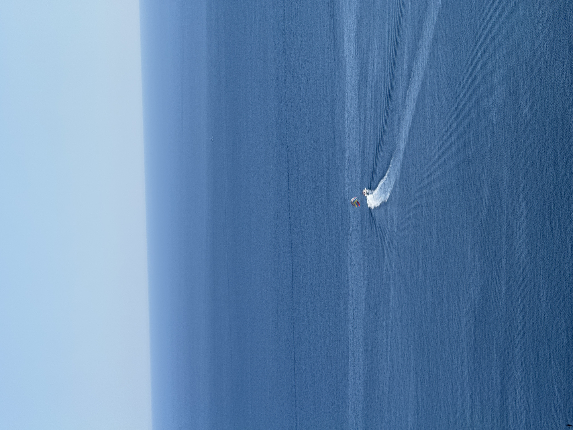 In the city of Seoce, Montenegro, a parasailer is being towed by a speed boat across the vast blue ocean. The parasailer is hoisted high in the air, a colorful canopy against the bright sky. The speed boat, leaving a white wake, is further out in the water, its engine working to pull the parasailer. The ocean is calm with gentle ripples, extending to the horizon where it meets the clear blue sky. The scene is serene and captures a moment of recreational activity on the water.