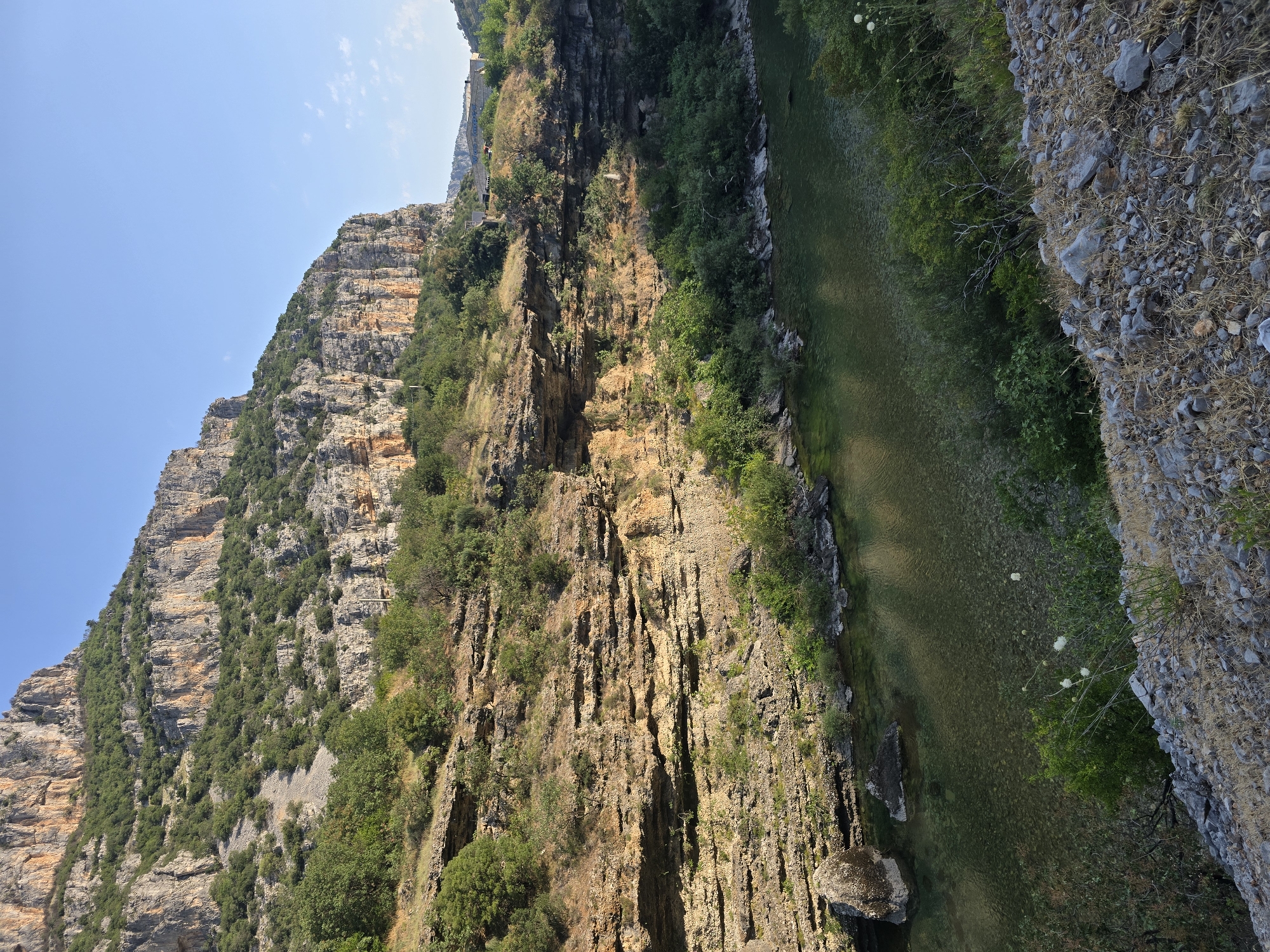 An elevated, vertical medium shot captures a sunlit canyon with a river flowing through it. The canyon walls, composed of layered rock formations, are rugged and textured, displaying hues of brown, tan, and orange. Lush green vegetation, including trees and shrubs, thrives on the slopes and near the riverbank. The river itself is clear and shallow, with visible pebbles on its bed. In the distance, buildings are nestled among the trees on a higher plateau, and a clear blue sky dominates the upper portion of the frame. The foreground consists of a rocky, barren slope with scattered dry grass and small white flowers. There are no people or animals visible in the scene.