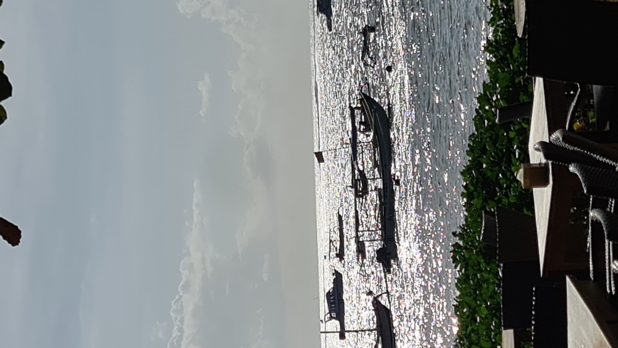 The image depicts a view of a beach in Denpasar, Indonesia, with several boats anchored in the sparkling, sun-drenched water. In the foreground, tables and chairs of an outdoor seating area are visible, suggesting a restaurant or cafe situated near the coast. Lush green foliage lines the edge of the seating area, bordering the beach. The sky above is partly cloudy, with the sun creating a brilliant, reflective surface on the ocean. The overall scene evokes a serene and picturesque coastal environment.