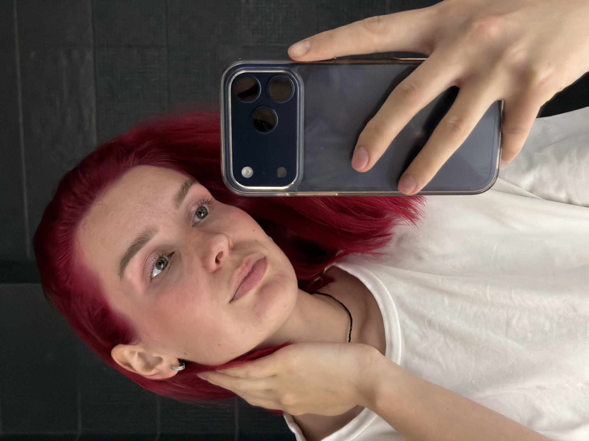 A person with red hair is holding a smartphone and taking a selfie. The person is wearing a white t-shirt. The background appears to be a dark surface, possibly tiles. There is no information to confirm the location is Chaikovsky, Russia.