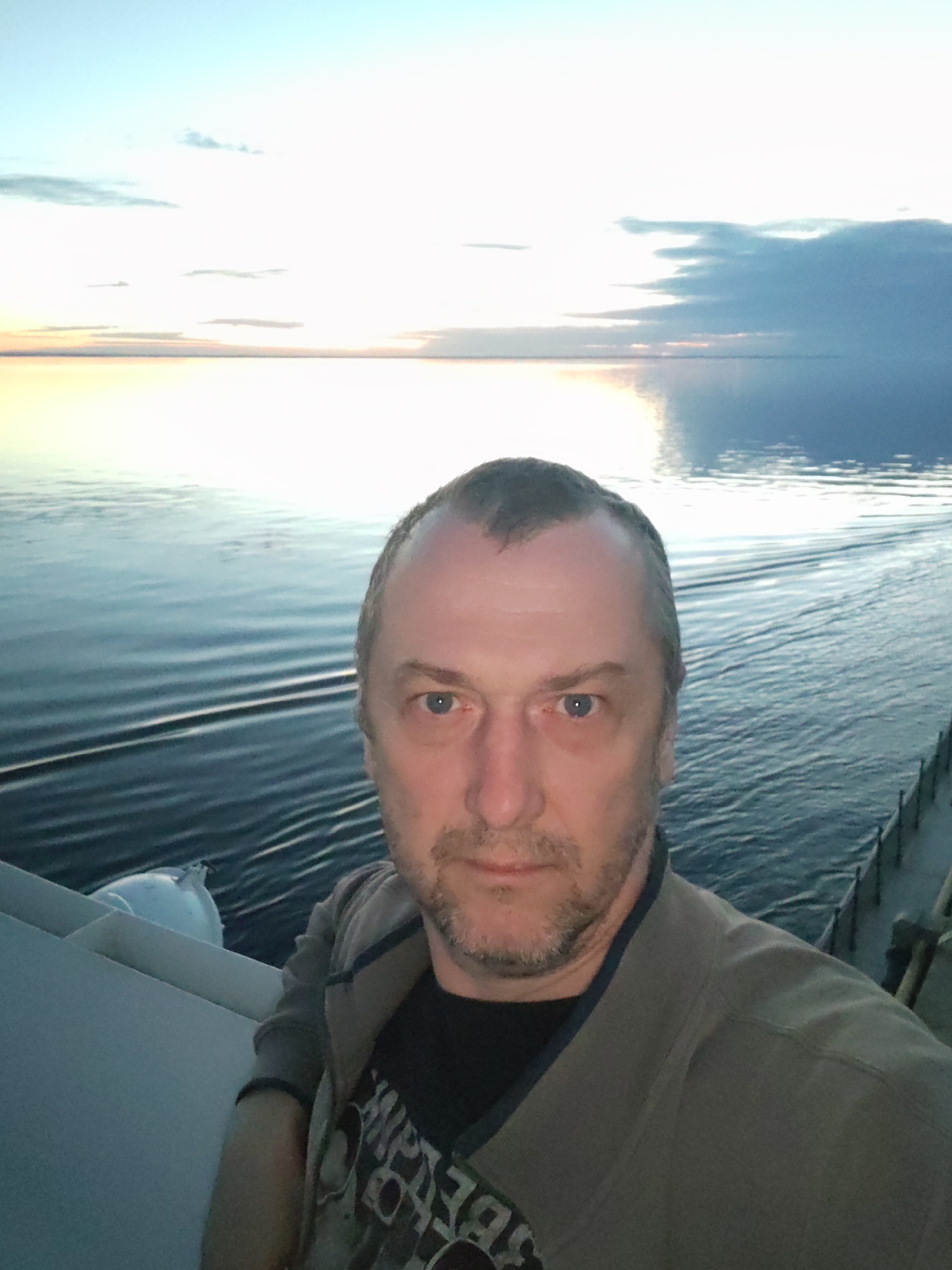 A male individual is featured in a selfie, positioned slightly to the left of the frame. He has light skin, blue eyes, a receding hairline with short dark hair, and a short beard. He wears a black t-shirt with stylized, unreadable white text on it, layered under an olive-green or khaki zip-up jacket. His gaze is directed towards the camera, with a neutral expression.
The man is standing on a boat or ship, with the deck railing visible on both sides. On the left, a white cylindrical fender or buoy rests on the railing. On the right, a metal railing with stanchions and lines is visible.
Behind the man, a large body of water, likely a sea or ocean, stretches to the horizon. The water on the right side of the frame shows distinct wake patterns, indicating the vessel is in motion. The horizon line is clearly defined, separating the water from the sky. The sky above is a blend of pale blue and white, with scattered clouds. Near the horizon, there's a soft, warm glow of yellow and orange, suggesting either sunrise or sunset. A darker cloud formation is visible on the far right of the sky. The overall lighting is soft and diffused, characteristic of twilight. The scene depicts a tranquil moment of travel on the water, potentially in the Russian location context provided.