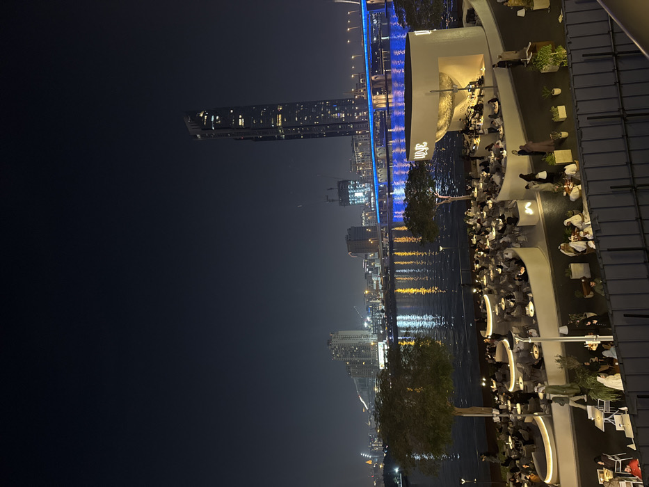 A night scene captures a city's edge with a blend of urban and social elements. A tall skyscraper pierces the dark sky, joined by other illuminated buildings. A body of water reflects the city lights, adding depth to the image. A terrace-like structure is filled with people at tables, suggesting a vibrant nightlife. A blue-lit bridge stretches across the scene, creating a striking visual element against the dark backdrop.