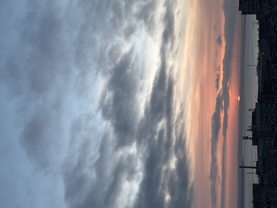 The scene captures an elevated panoramic view of Burgas, Bulgaria, at either sunrise or sunset, overlooking the city and the Black Sea. The sky exhibits a layered cloud cover, transitioning from pale blue and grey at the zenith to vibrant orange, pink, and yellow hues near the horizon. A distinct, partial disc of the sun, glowing orange-yellow, is visible directly above the water surface towards the central-right of the frame. Multiple dark, avian silhouettes are discernible against the illuminated sky and water, primarily clustered near the sun. The foreground and midground consist of the upper portions and rooftops of numerous multi-story urban buildings, rendered in silhouette. Several communication antennas are mounted on the rooftops. A tall, dark industrial-type stack or chimney is prominent on the left side of the urban silhouette. No human subjects are present.