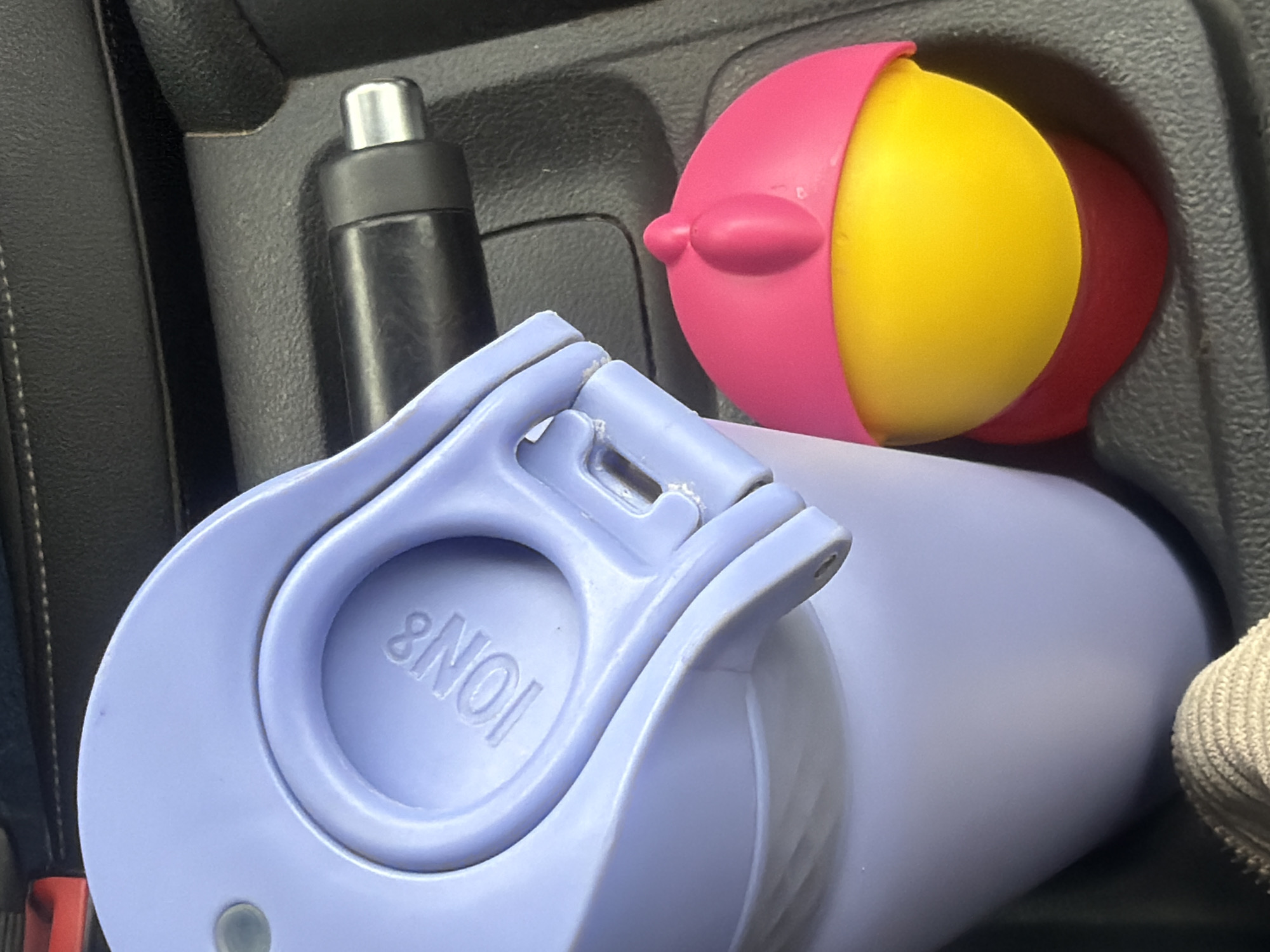 The image shows the interior of a car. In the center console, there is a black travel mug with a silver lid, a large, light purple water bottle with an "ION8" brand marking on the lid, and a toy ball. The toy ball is segmented into pink, yellow, and red sections. The car interior appears to be dark. The image was not taken in Kamburovo, Bulgaria.