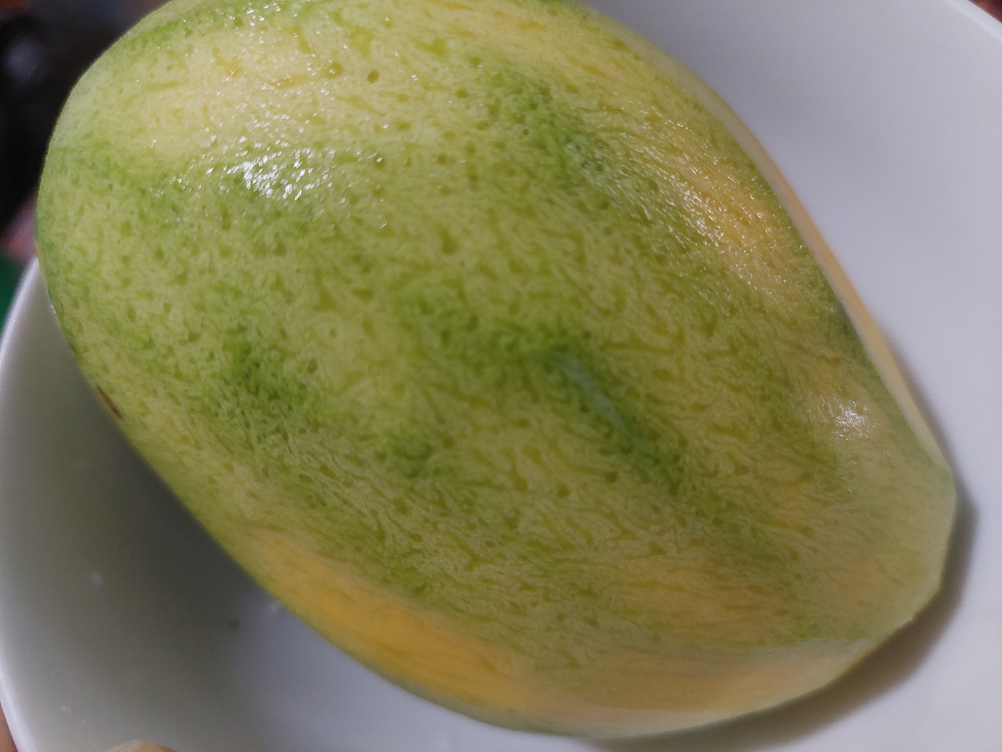 A close-up, overhead shot shows a partially ripe mango with streaks of yellow and green skin resting in a white bowl. The mango is oval-shaped and smooth, with a slightly textured surface. The background is blurred, but appears to be a white surface, possibly a table. The lighting is bright, creating highlights on the mango's skin. This image does not contain any identifiable features related to the city of Jelutung, Indonesia.