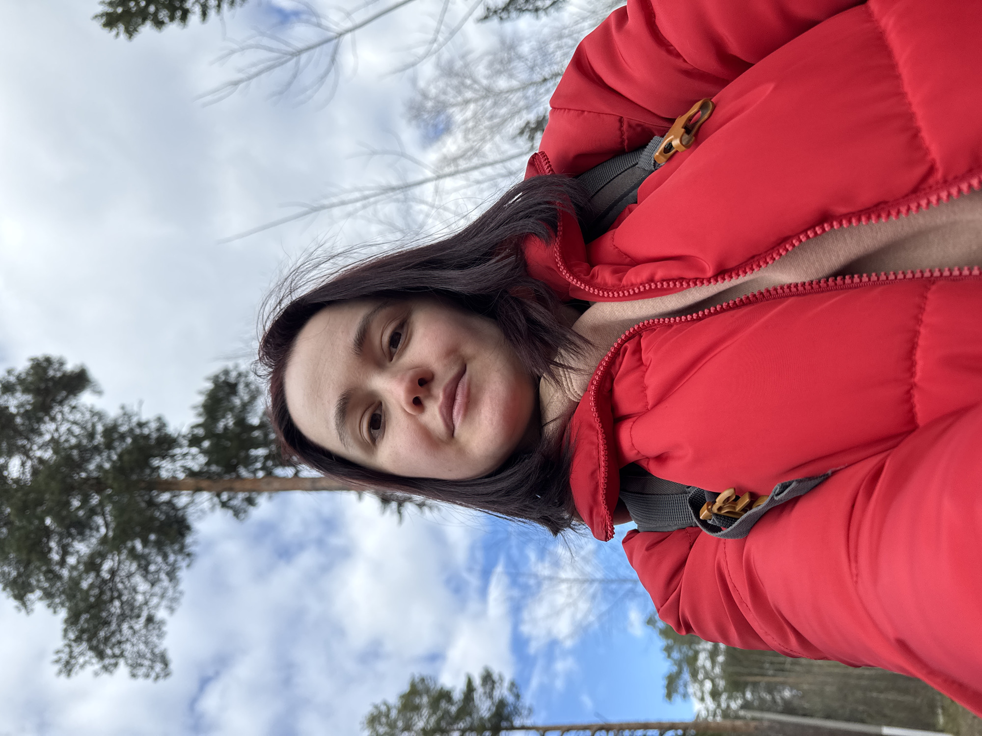 A woman is taking a selfie outdoors in what appears to be a park or wooded area. She is wearing a red puffer jacket and has dark hair. The sky is visible in the background with scattered clouds and trees. Although there are trees in the background, the location within Izhevsk is not identifiable.