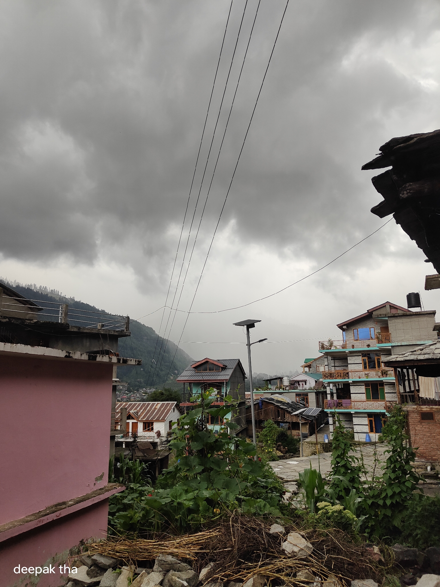 The image shows a view of a town in Vashisht, India, on a cloudy day. The foreground features a pile of stones and dried branches. To the left, a pink-colored building is partially visible. Beyond this, the scene opens up to a cluster of multi-story buildings with colorful balconies and windows, characteristic of the region. A road or pathway winds through the town. In the background, a lush green mountain slope rises, covered with trees and houses. Power lines are strung across the sky, which is dominated by heavy, gray clouds, suggesting impending rain. A solar-powered street light stands near a group of buildings.