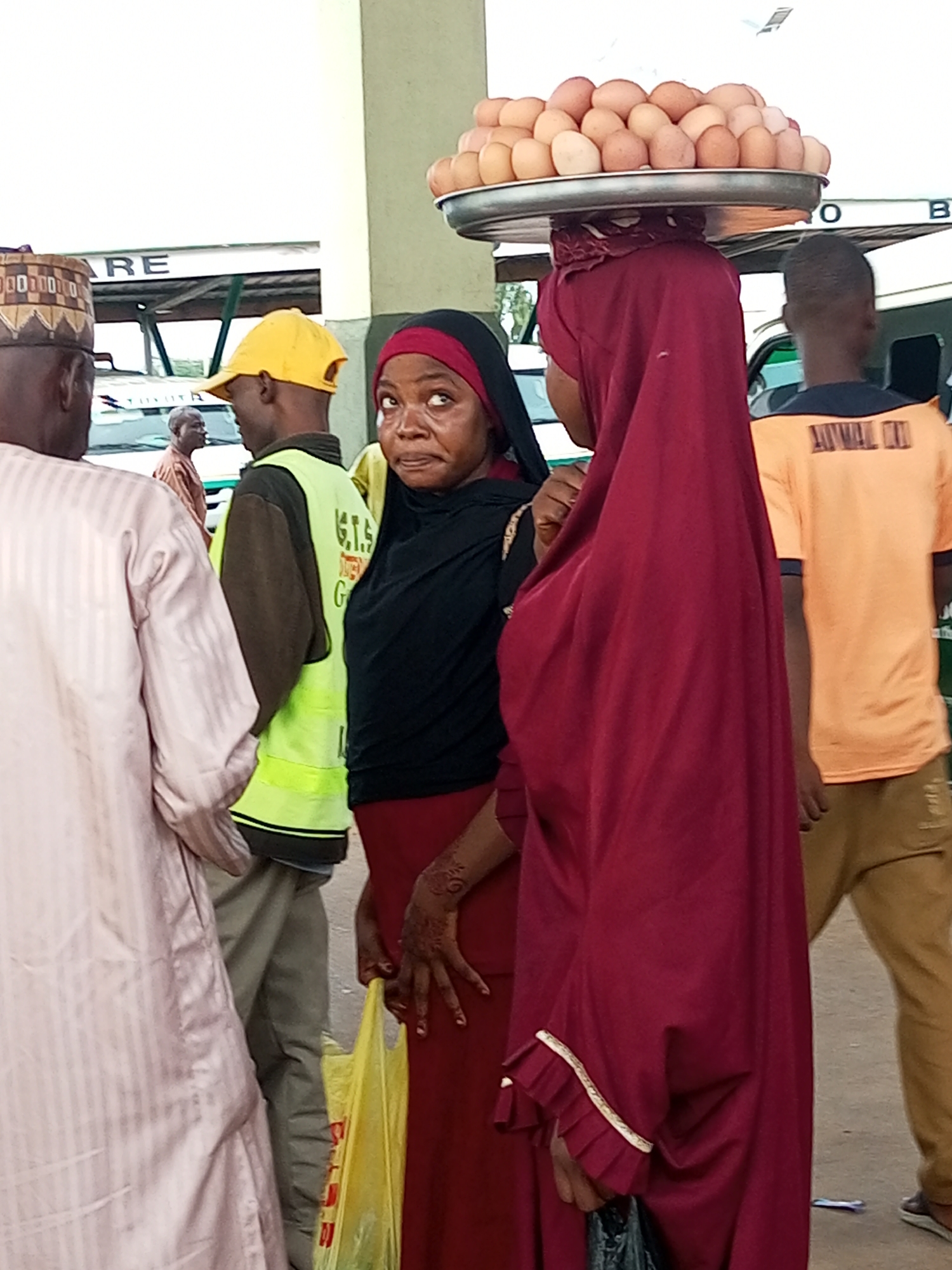 A group of people, including women with hijabs and men in traditional attire, are seen in an outdoor setting that appears to be a market or transportation hub in Gombe, Nigeria. One woman in the foreground, wearing a dark hijab and a red dress, has a tray of eggs balanced on her head. Her face shows a concerned expression as she looks upwards and to the left. Next to her, another woman in a maroon hijab stands with her back to the camera, partially obscuring the woman with the eggs. To the left of the group, a man in a light-colored, striped robe is visible from the side, facing left. Another man in a bright yellow cap and a neon green vest is positioned behind him, facing towards the viewer. In the background, there are structures with signage that includes the word "RE" and other illegible text, suggesting a commercial or transport area. A person wearing an orange t-shirt is also visible in the background on the right side of the image. The ground is paved, and the overall lighting suggests it is daytime.