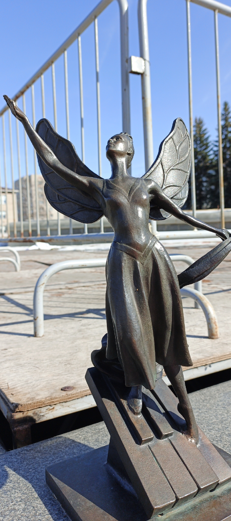 A full-shot photograph shows a bronze statue of a winged woman on a bridge in the city of Chaikovsky, Russia. The statue is positioned so that her left hand is raised in the air. She appears to be wearing a dress that reaches to her knees, and she stands atop a bronze base that resembles a piano keyboard. The background includes a silver railing, a wooden deck, and some buildings and trees under a clear blue sky. The sun casts shadows on the deck and the statue's base. The statue has a patina that gives it a dark, almost brown appearance.