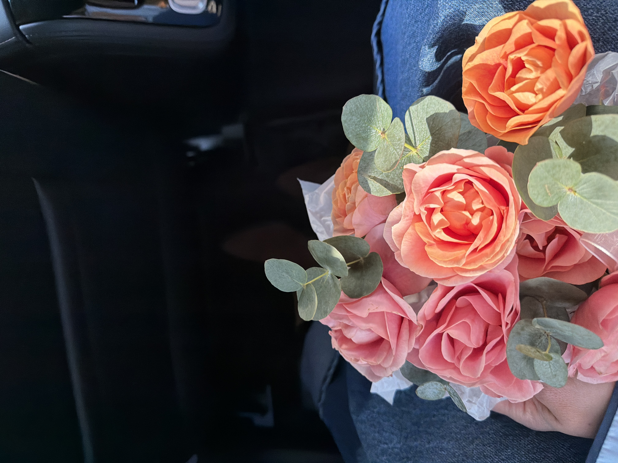 A person is seated inside a vehicle, holding a bouquet of flowers on their lap. The bouquet consists primarily of light pink and orange roses, interspersed with green eucalyptus leaves and white tissue paper. The hand holding the bouquet, visible on the lower left, has light skin tone. The person is wearing blue denim jeans, partially visible on their lap. The background shows the dark interior of a car, specifically the dashboard or passenger footwell area, with various dark gray and black surfaces and faint reflections of interior components. Sunlight illuminates the flowers and denim, casting distinct shadows. The specific location within the city of Kukui, Russia, cannot be determined from the visual information provided in the image.