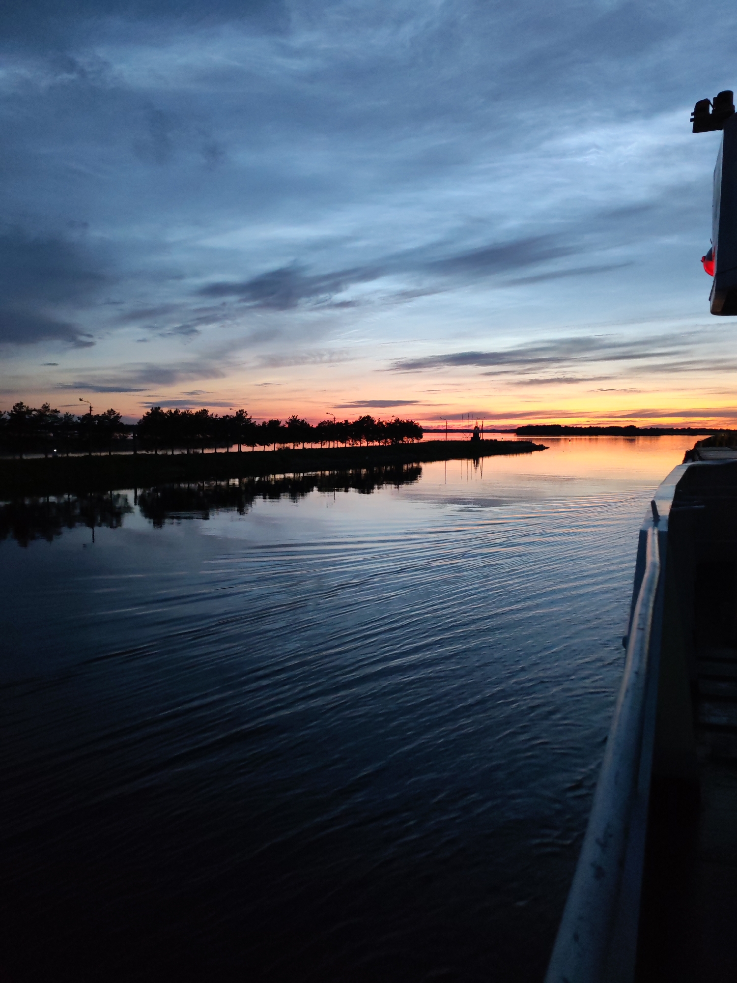 The image depicts a sunset over a body of water, possibly a river or lake, with a shoreline lined with trees. The sky is filled with dramatic clouds painted in hues of blue, grey, and orange, indicating the last moments of daylight. The water reflects the sky and the treeline, creating a mirror image. In the foreground, the edge of a boat or vessel is visible on the right side of the frame. The water exhibits ripples, suggesting movement. On the distant shore, a few structures, including what appears to be a crane or industrial equipment, are silhouetted against the sunset. The overall atmosphere is serene and tranquil. The location is identified as the city of Sheksna, Russia.