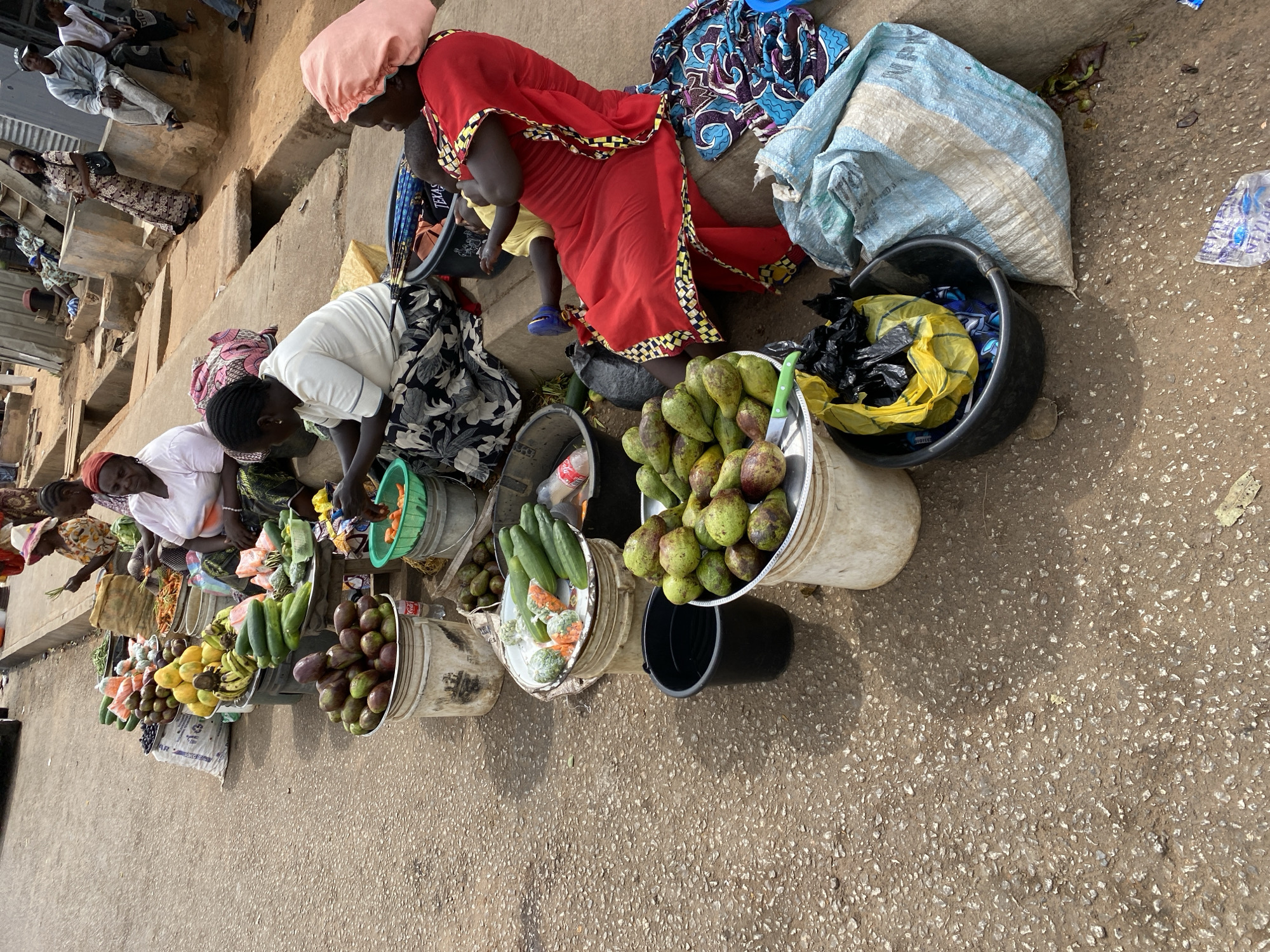 This overhead shot captures a vibrant outdoor market scene in Bukuru, Nigeria. The image is dominated by several women vendors engaged in selling fresh produce, displayed directly on the ground in various buckets and metal basins.
In the mid-ground, a woman wearing a striking red dress with yellow and black trim and a pink headscarf is crouched, seemingly attending to a small child in yellow and blue shoes who is partially visible beside her. To her right, a large assortment of green and reddish avocados is piled high in a white bucket, with a green knife resting on a nearby tray. Another woman, dressed in a white top and a dark patterned skirt, is seen leaning over her display of produce, which includes more avocados, bananas, and papayas. Further down the line, other women are also attending to their stalls, featuring items like cucumbers and what appear to be carrots or similar vegetables in a green plastic bucket.
The produce is neatly arranged in a line along the ground. In the background, more people are visible, some sitting on raised concrete steps or platforms, suggesting a busy public space. The ground is a light-colored, pebbled surface.
Visible text includes "TEXAS" on a blue and white patterned fabric bag near the woman in red, "WPM" on a large light blue woven sack, and "DIGIEES" or similar on a black bag inside a black bucket. A mini Coca-Cola bottle can also be seen among the produce. The scene is brightly lit, indicating it is daytime, and the overall impression is one of active commerce in a local market.