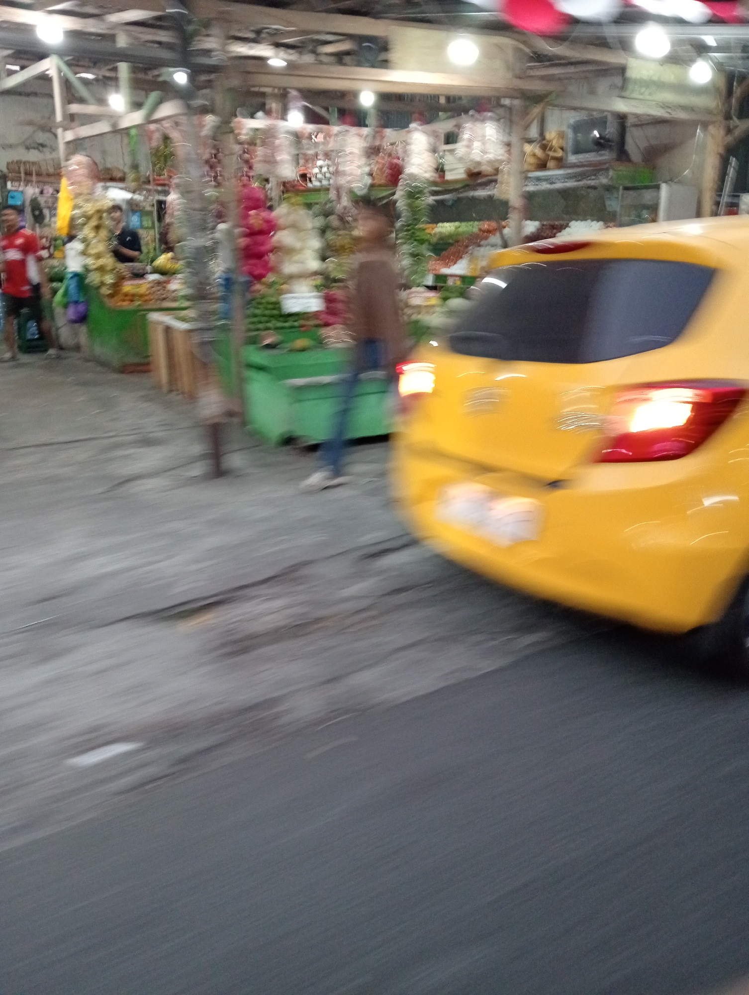 The image depicts a blurry outdoor scene, likely a market or roadside stall area, in Ngaglik, Indonesia. A yellow car is the most prominent subject, with its rear end facing the camera and its taillights illuminated. The car is positioned on a paved road, which occupies the lower portion of the frame. In the background, market stalls are visible, featuring produce such as fruits and vegetables. Several people are present, though their details are obscured by motion blur. One person in a red shirt is standing near a green stall on the left, while another figure in brown is walking in front of the stalls. The environment appears to be dimly lit, possibly indicating evening or an overcast day, with overhead lights illuminating the market area. The overall impression is of a busy, dynamic scene captured with a fast shutter speed.