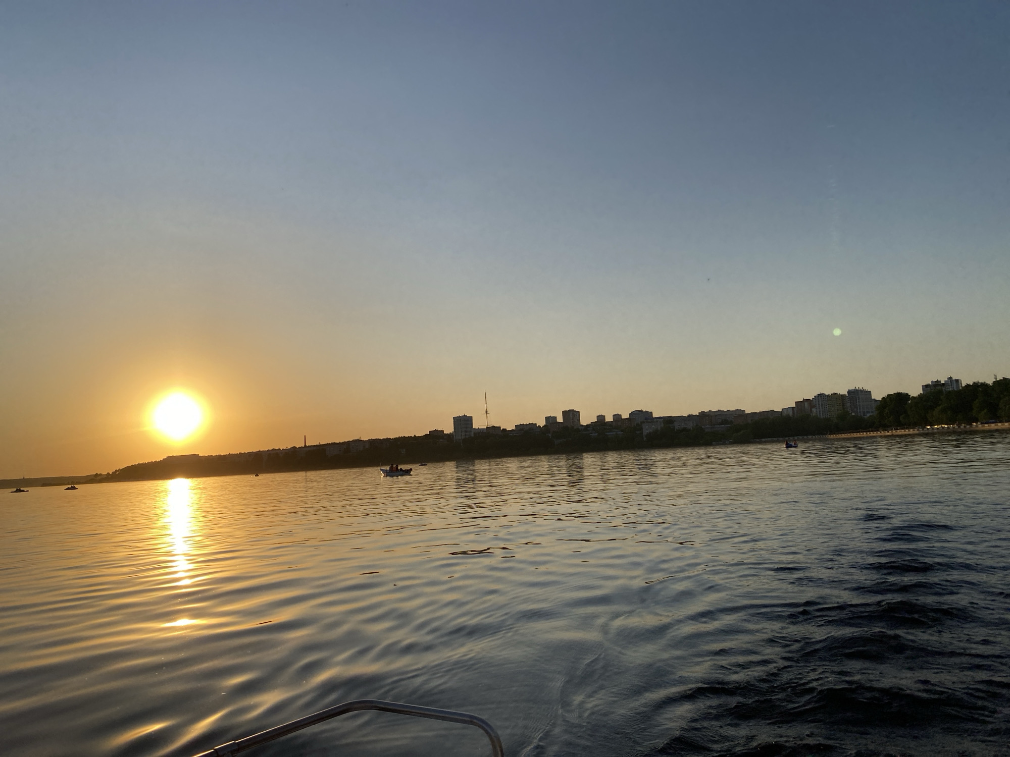 The image captures a sunset over a body of water, likely a reservoir, in Izhevsk, Russia. The sun is positioned low on the horizon on the left side of the frame, casting a golden reflection on the water's surface. The water is visibly rippled, indicative of a slight breeze or current. On the opposite bank, a cityscape silhouette is visible, comprised of buildings of varying heights and interspersed with trees. In the water, there are a few small boats, indicating recreational activity. The sky transitions from a golden hue near the horizon to a light blue higher up. The bottom of the image includes part of a metal boat rail, suggesting the photo was taken from a boat.