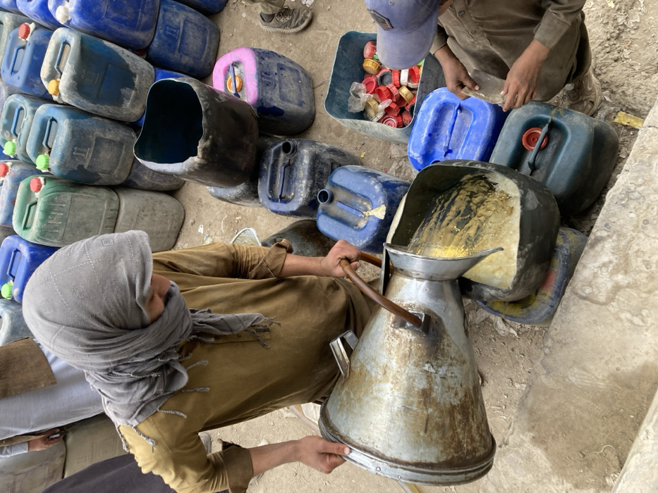 This image depicts a scene of liquid transfer in Quetta City Tehsil, Pakistan. A person, wearing a brown tunic and a gray headscarf covering the head and lower face, holds a large, tarnished metal funnel-shaped container with a wooden handle, tilting it to pour a yellowish, viscous liquid into an open, dark-colored plastic jerrycan. To the lower right, a second individual, wearing a light blue baseball cap and brown clothing, crouches near a plastic bin containing numerous red, yellow, and white plastic caps, handling small, clear plastic pieces. The ground is uneven dirt. In the background and surrounding the figures, dozens of plastic jerrycans, mostly blue and green, are stacked and scattered, many showing signs of wear and dirt. A third individual is partially visible in the far left background.