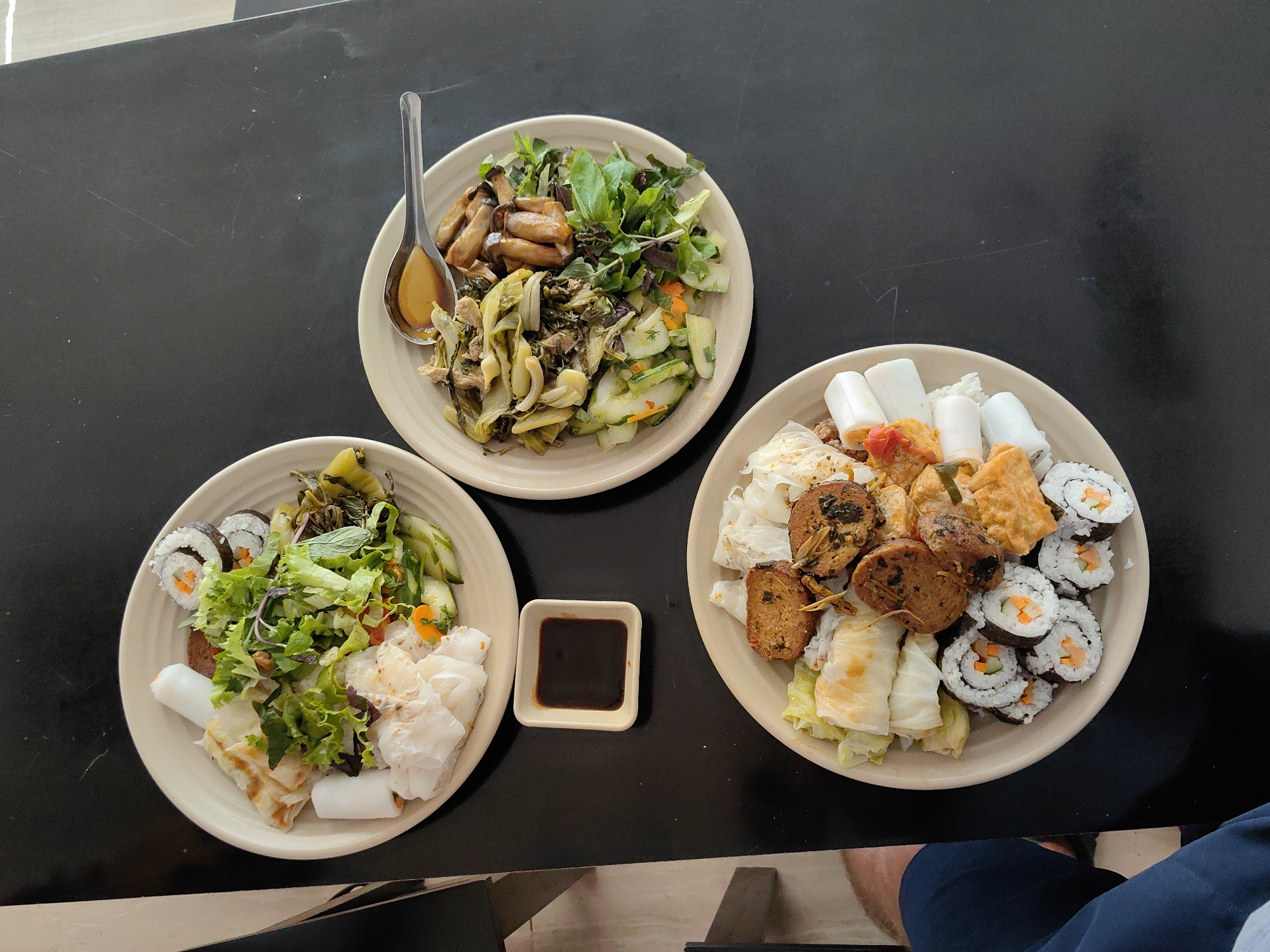 Three plates of food are arranged on a dark, likely black, table. The setting is indoors, possibly a restaurant, inferred from the presentation of the food.
The plates appear to contain a mix of Asian dishes, likely Vietnamese based on the presence of rice paper rolls and fresh herbs. One plate has stir-fried vegetables, mushrooms, and fresh greens. Another contains sushi rolls, rice paper rolls, salad, and other unidentified items. The third plate contains rice paper rolls, deep-fried items, and sushi. A small square bowl containing a dark liquid (likely soy sauce or a similar condiment) sits between two of the plates.
Partial view of a person in dark blue is visible in the lower right.
The image metadata indicates this was taken in Hong Gai Ward, Vietnam.