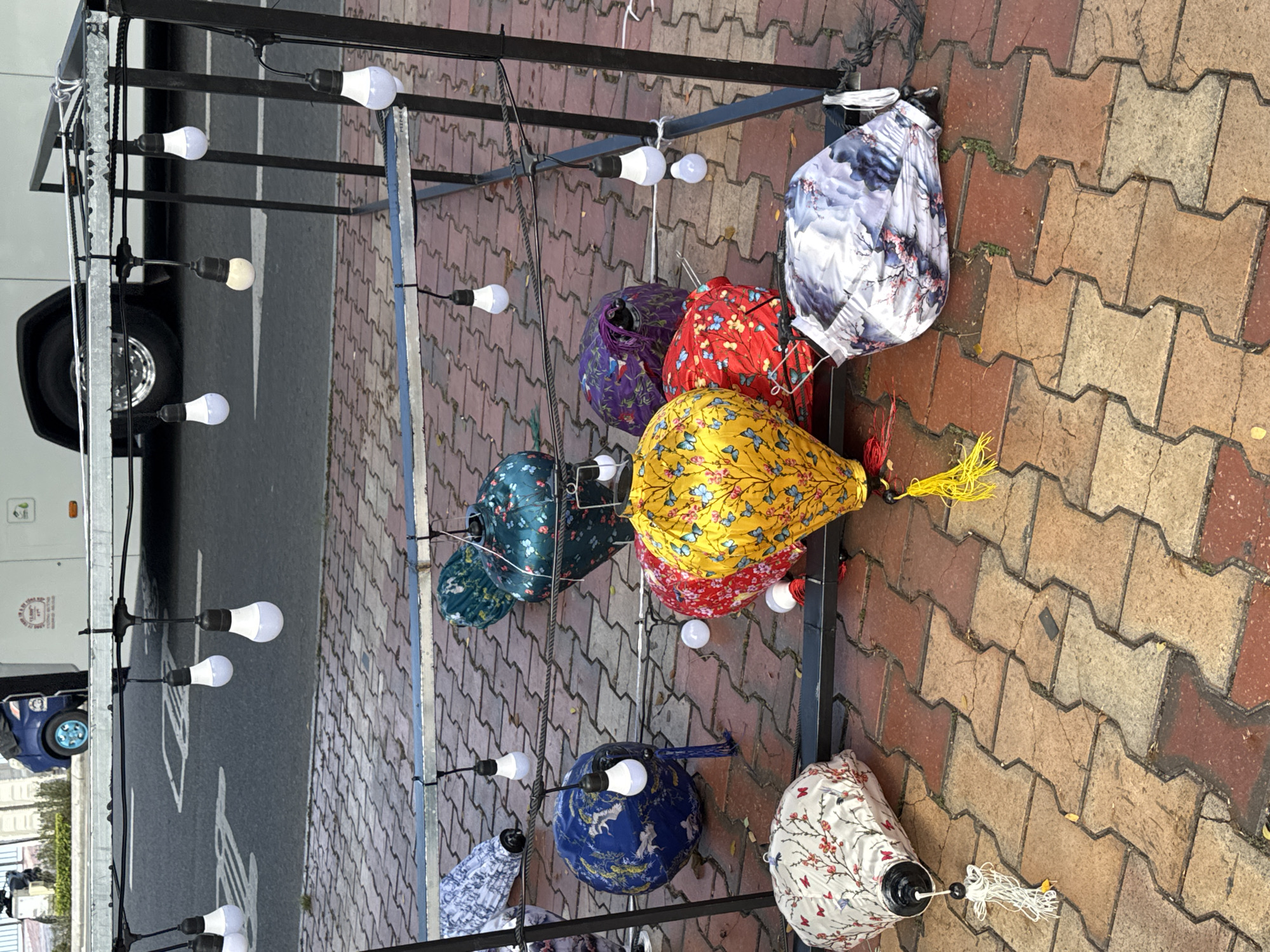 This image depicts an outdoor display of lanterns and string lights, likely set up for an event or market, situated on a sidewalk paved with interlocking bricks. In the background, a white bus is visible, and a blue vehicle with a distinctive teal-colored wheel is parked on the street. The street itself has white lane markings, suggesting it is a public road. The lanterns are of various designs and colors, including floral patterns in red, yellow, and green, as well as a blue lantern with a detailed depiction of animals and a white lantern with a cherry blossom motif. Some of these lanterns are hanging from a metal structure, while others are placed on the ground. String lights with white LED bulbs are also suspended above and around the lanterns, some of which are lit, contributing to the festive atmosphere. The scene appears to be in a city environment, and considering the context of the lanterns and the bus, it is plausible that this scene is located in Đà Nẵng, Vietnam, a city known for its vibrant cultural events and displays.