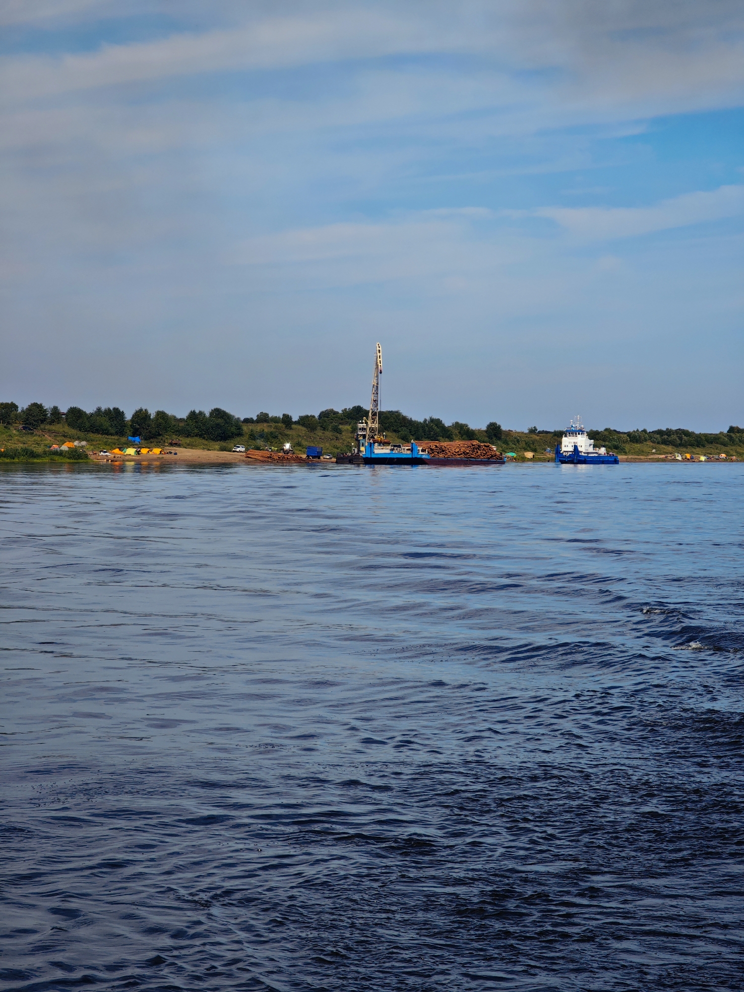 This image depicts a wide river with a calm surface, showing gentle ripples and waves. In the background, a sandy shore is lined with green trees and shrubs. On the shore, there are several orange and yellow tents, suggesting a campsite or temporary settlement. A large blue crane is positioned on a barge, which is loaded with a significant amount of logs. A blue and white boat is anchored near the shore. The sky above is a pale blue with scattered white clouds. The scene appears to be a daylight setting, possibly in a rural or semi-industrial area along a waterway. The provided location is the city of Yarcevo, Russia, suggesting this river could be part of the Dnieper River or one of its tributaries. There are no visible people in the image.