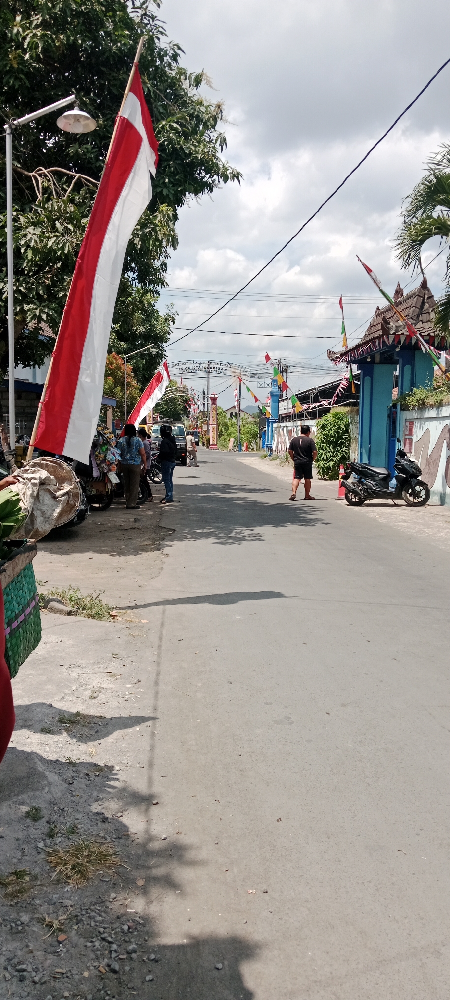 The image depicts a street scene in Purwomartani, Indonesia, likely during a festive occasion given the prominent display of Indonesian flags (red and white banners) and smaller decorative flags. In the foreground, on the left side of the street, there is a stack of large, wrapped objects next to a woven green basket, possibly containing produce. Several people are gathered near a parked motorcycle, some standing and observing the surroundings. A lamppost with a visible bulb stands to the left. The street itself is paved and appears to be a public thoroughfare. Further down the street, a blue building with decorative elements and a traditional roofline is visible, adorned with more flags. A black scooter is parked in front of this building. A person is seen walking away from the camera on the right side of the street. The environment is characterized by trees, power lines, and a cloudy sky. The overall impression is of a community preparing for or celebrating an event.