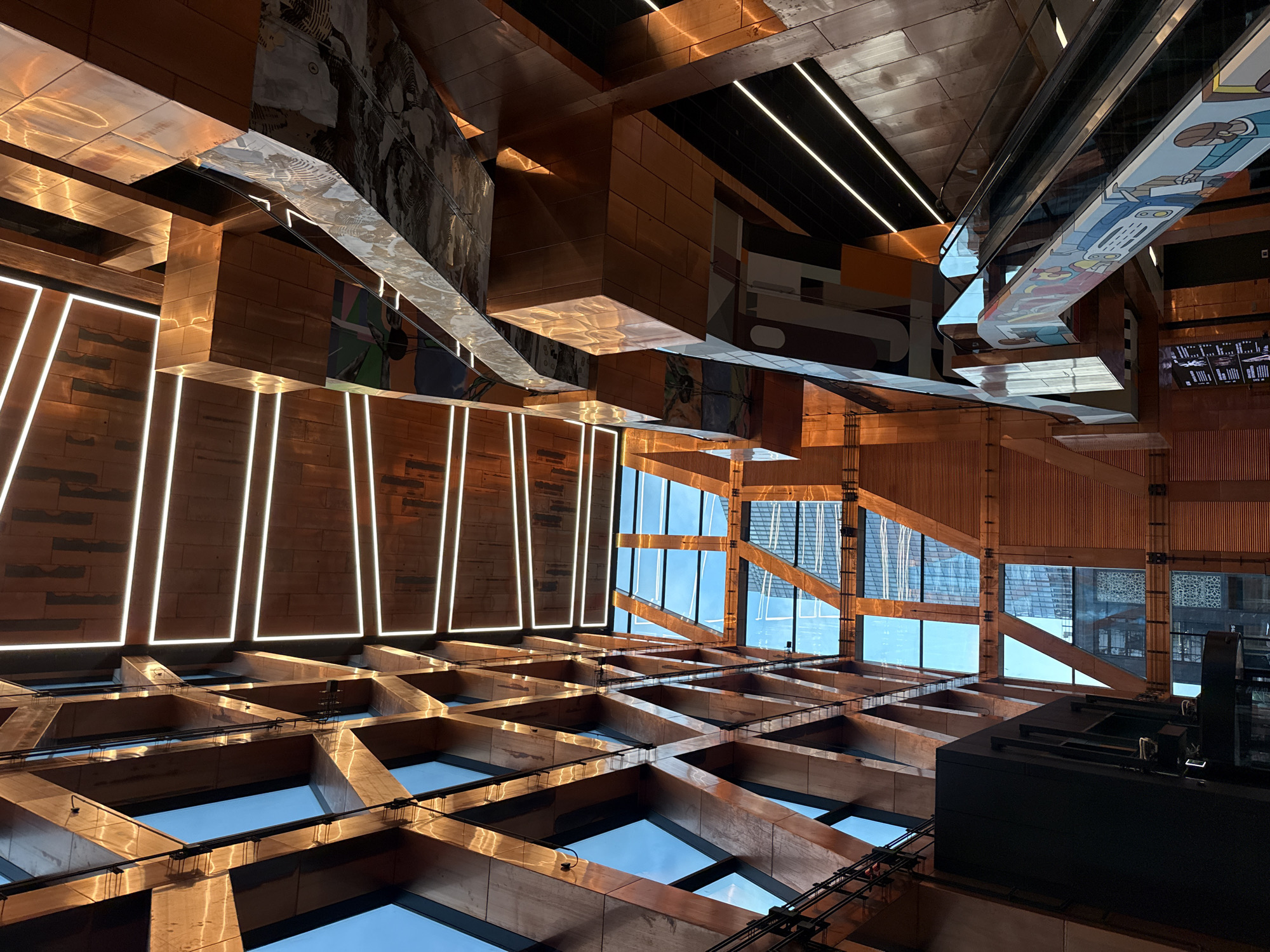 An interior shot captures a modern architectural space characterized by a striking interplay of copper-toned metallic surfaces and geometric design. The perspective is from below, looking upwards, emphasizing the height and complexity of the structure. A large section of the left side of the image is dominated by a crisscrossing wooden lattice structure with rectangular glass panels, allowing a glimpse of a blue sky and distant buildings.
To the right, the architecture becomes more angular and layered, with protruding geometric forms clad in the same copper-like material. These surfaces are highly reflective, catching light and creating warm, metallic glints. Linear white LED lights are strategically placed along the edges of some of these copper elements, accentuating their forms and adding a contemporary touch.
In the middle ground, a large, multi-level structure with curved, metallic surfaces is visible. This area is adorned with murals or graphic designs, featuring bold colors and abstract patterns, some appearing to depict figures or scenes. Below this, an escalator is visible, its sides decorated with colorful illustrations that appear to be cartoonish figures and objects. The escalator treads are dark and reflective.
Further into the background, through the open spaces of the architecture, more distant building facades are visible, hinting at an urban environment beyond the interior. The lighting in the space appears to be a combination of natural light from the windows and artificial lighting from the embedded LEDs. There are no people clearly visible in the shot, focusing solely on the architectural elements and their aesthetic qualities.