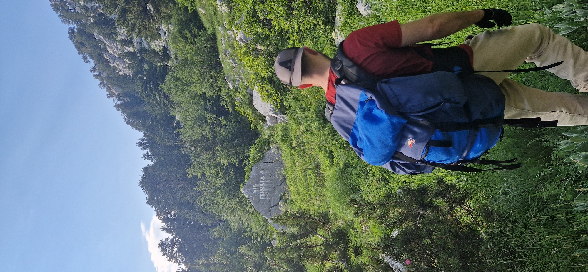 A person wearing a red shirt, tan pants, a grey baseball cap with an "A" on it, and black gloves is walking away from the camera. They are carrying a large blue and grey backpack. The person is in a grassy, mountainous area with trees and rocks. A large rock with "VIA FERRATA" and an arrow pointing to the right is visible in the background. The sky is blue with some clouds. This is the location of Kruševice, Montenegro.