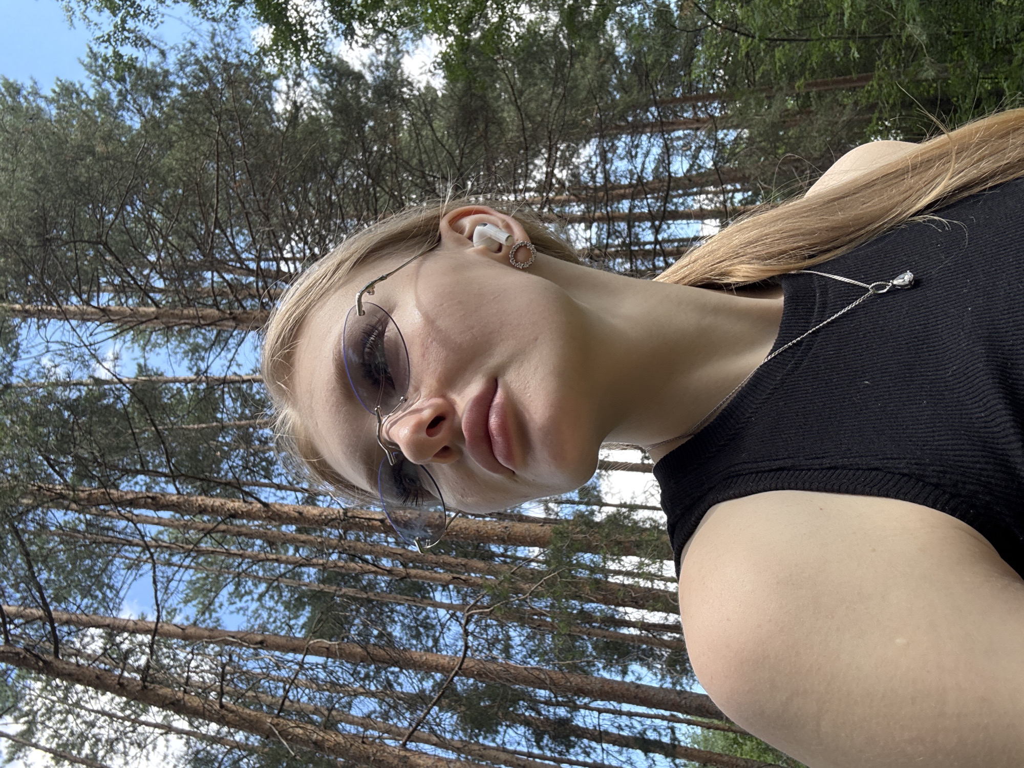 A young woman with blonde hair, wearing a black tank top, sunglasses, and earrings, stands in a forest. She has an earbud in her right ear and a necklace with a pendant. The trees in the background are tall and thin, with green foliage visible against a blue sky with scattered clouds. The lighting suggests it is daytime. The location is a forest in Perm, Russia.