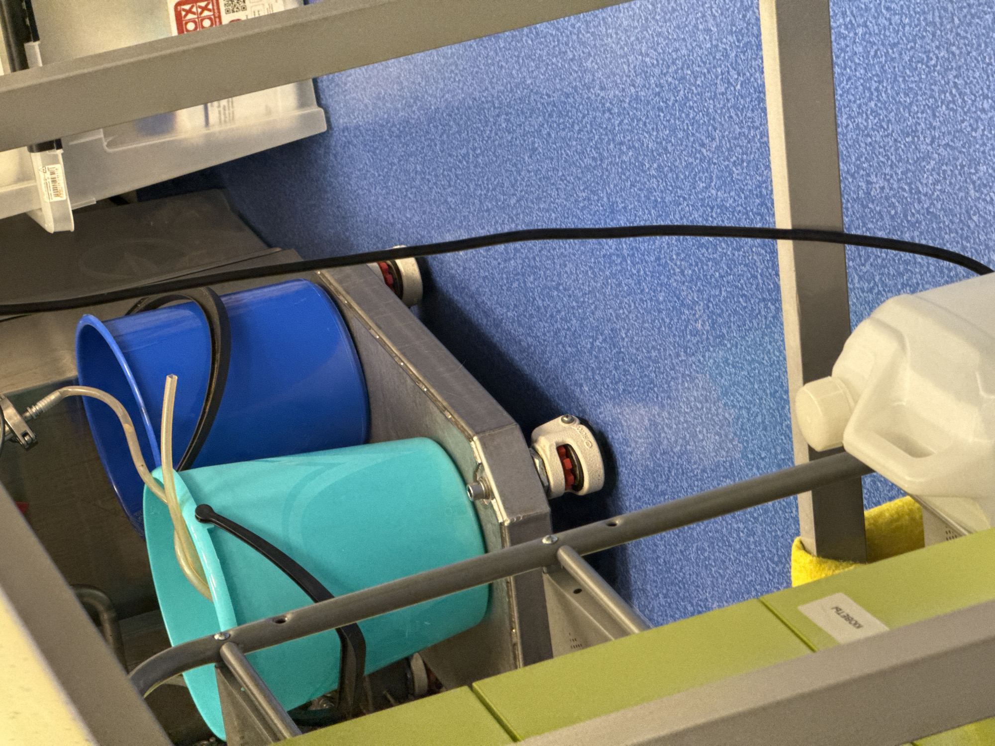 This image depicts an indoor scene within what appears to be a utility area, laboratory, or storage space, characterized by functional shelving and various containers. The background features a textured blue wall.
In the midground, a robust metal shelving unit dominates the lower and central parts of the frame. On a lower platform or shelf, two plastic buckets are arranged: one is bright blue, and the other is a vibrant turquoise. Clear tubing, suggesting a system for liquid transfer or filling, is visible extending into and around the turquoise bucket, with some connections to the blue one. A black cable stretches horizontally across the blue wall, passing through the metallic framework.
On a higher shelf to the right, a white plastic jug with a handle is partially visible. Above the buckets, on an upper shelf of the metal unit, rests a clear plastic storage bin. This bin features several labels, including a white one with a black barcode and unreadable text, and a red label displaying white symbols resembling two overlapping 'X's, possibly indicating a warning or classification. A QR code is also present on this label.
Further to the right, a yellow sponge or cloth is tucked beneath a metal support beam. A flat, light green surface, likely a shelf or part of a workspace, is visible in the lower right corner, marked with a white sticker bearing the black alphanumeric text "P22800". The overall arrangement suggests an organized, practical setup for tasks involving liquids or general storage. The location context provided is Moscow, Russia.