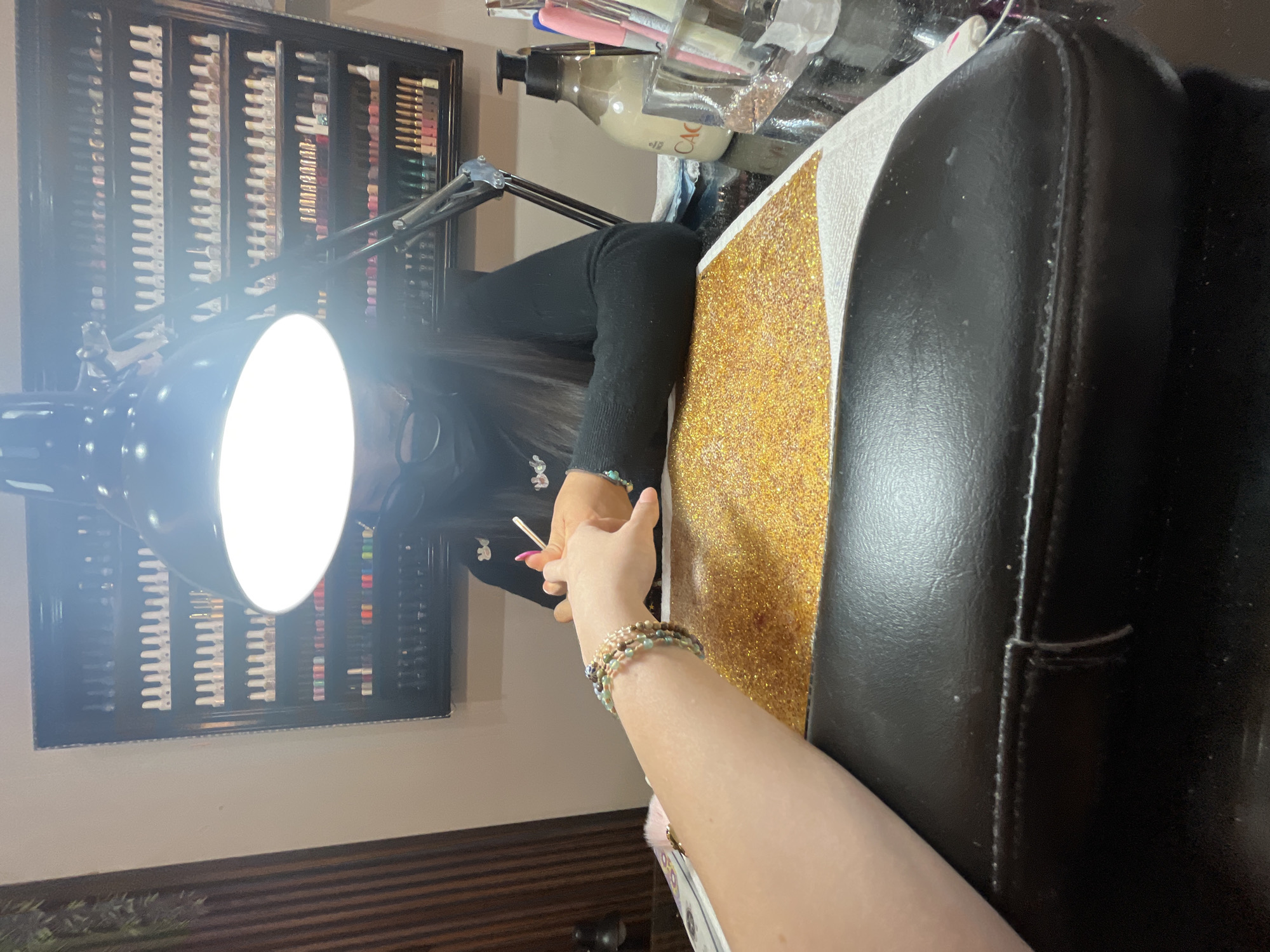 The image depicts an indoor scene within a nail salon in Puebla, Mexico. In the foreground, a black padded armrest is visible. The central focus features a client's left forearm and hand, adorned with multiple beaded bracelets, resting on a white mat partially covered with gold glitter. Directly opposite, a nail technician is seen partially obscured by a bright, adjustable desk lamp with a circular light, actively working on the client's hand with a small tool. The technician wears a black long-sleeved top and a dark face mask. To the right of the workspace, a pump bottle labeled "CAO" and a clear container holding various brushes and tools are present on a reflective black surface. The background features a large, dark display cabinet with multiple shelves, meticulously organized with numerous nail polish bottles of varying colors. The wall to the left exhibits vertical wooden paneling.