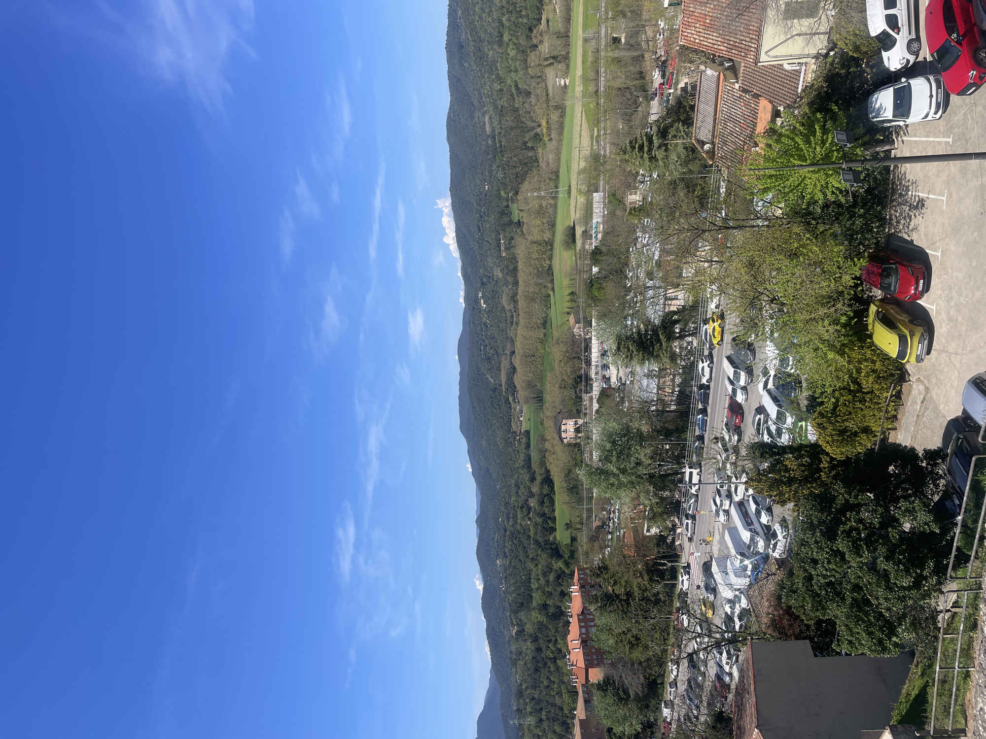 The image shows a landscape taken from an elevated position in Hostalric, Spain. The left side of the image primarily consists of a clear blue sky with wisps of white clouds.
In the center of the image, a mountain range is visible, covered in lush green vegetation. At the foot of the mountains, there is a flat green field, potentially a meadow or farmland.
On the right side of the image, a densely populated area is seen. There are numerous buildings with red-tiled roofs. A parking lot filled with various cars, predominantly white, is present. Trees and greenery are interspersed among the buildings and parking area.