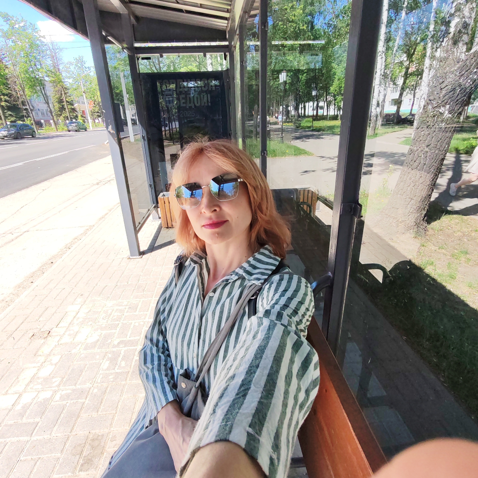 A woman with reddish-blonde hair, wearing sunglasses and a striped shirt, is sitting on a bench at a bus stop. The bus stop has a metal frame and glass panels. Through the glass, a street, trees, and a sidewalk can be seen. A person is walking on the sidewalk outside the bus stop. Cars can be seen on the road. The location is likely Chaikovsky, Russia, based on the architecture and environment.