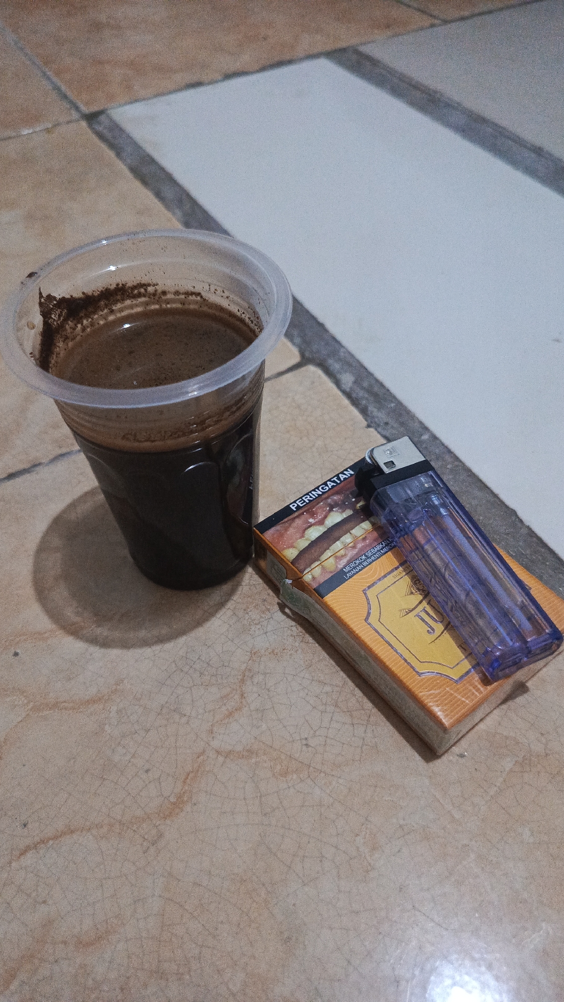 A close-up, overhead shot shows a plastic cup filled with dark liquid and coffee grounds, placed on a tiled surface. To the right of the cup is a pack of cigarettes with an orange and yellow design, featuring an image of a burger and the Indonesian word "PERINGATAN" (Warning). A transparent blue lighter rests on top of the cigarette pack. The tiled floor has a pattern of beige and white squares separated by dark grout lines, with a visible shadow cast by the cup on the lower left. The overall setting appears to be an indoor environment, possibly a home or a simple establishment, given the tiled flooring. There are no individuals present in the image. This scene is a common sight in various locations within Tangerang, Indonesia, from personal residences to small warungs or local eateries, where coffee and cigarettes are frequently consumed together.