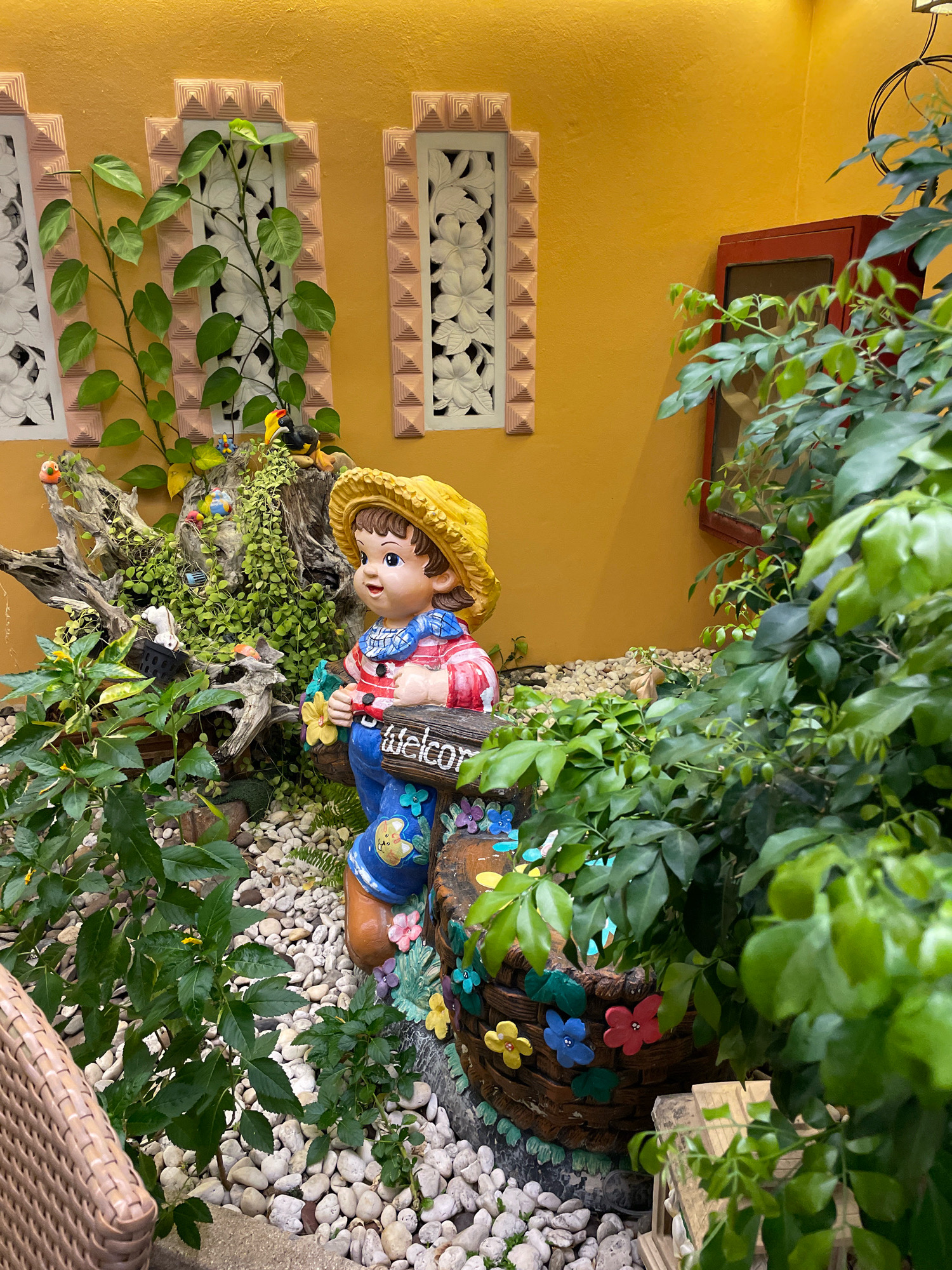 A colorful garden figurine of a child in a straw hat and blue overalls holding a "welcome" sign stands amidst lush green plants and white pebbles. The figurine is dressed in a striped shirt and has a friendly expression. Behind the figurine, a weathered piece of driftwood is adorned with small ceramic birds and cascades of green ivy. The background features a bright yellow wall with ornate window frames. To the right, a dense bush of green foliage partially obscures a red-framed cabinet. The scene appears to be a decorative outdoor space, possibly in a cafe or restaurant, given the cheerful aesthetic. The image's composition and style suggest it could be located in Pattaya City, Thailand, known for its vibrant and tropical garden designs.