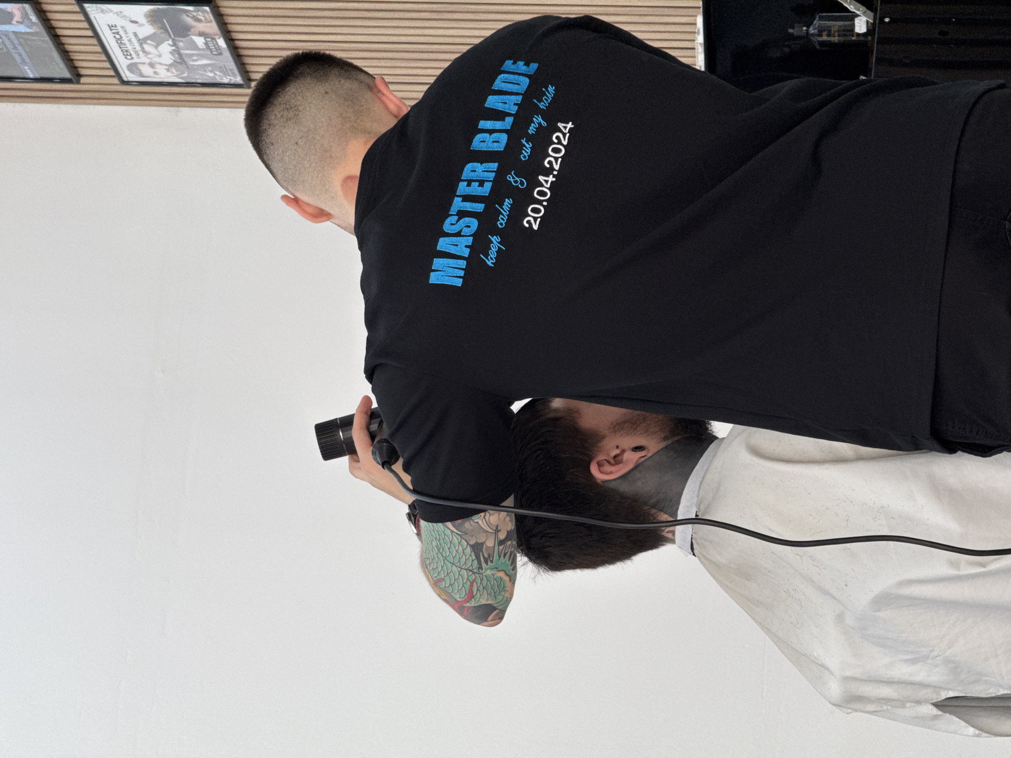 The image depicts two men in what appears to be a barber shop or salon in Sofia, Bulgaria. One man, seen from behind, is wearing a black long-sleeved shirt and holding a camera, seemingly taking a photograph. His left arm reveals a prominent, colorful tattoo featuring a dragon motif. The black shirt has text printed in blue: "MASTER BLADE" in large, bold letters, followed by "keep calm & cut my hair" in a smaller, cursive font, and the date "20.04.2024."
The second man is seated below and slightly in front of the photographer, viewed from an overhead angle. His head is tilted downwards, and he is draped in a white barber's cape which is speckled with small hair trimmings, indicating a recent haircut. He has dark hair and a beard, and black tunnel earrings are visible in his ears. A black cable runs across the white cape, connected to an unseen device.
In the background, mounted on a light brown, slatted wall, are framed items, one clearly displaying the word "CERTIFICATE." To the far right, part of a barber's station or mirror with various grooming tools can be glimpsed. The scene suggests a post-haircut photoshoot or documentation within a professional grooming environment.