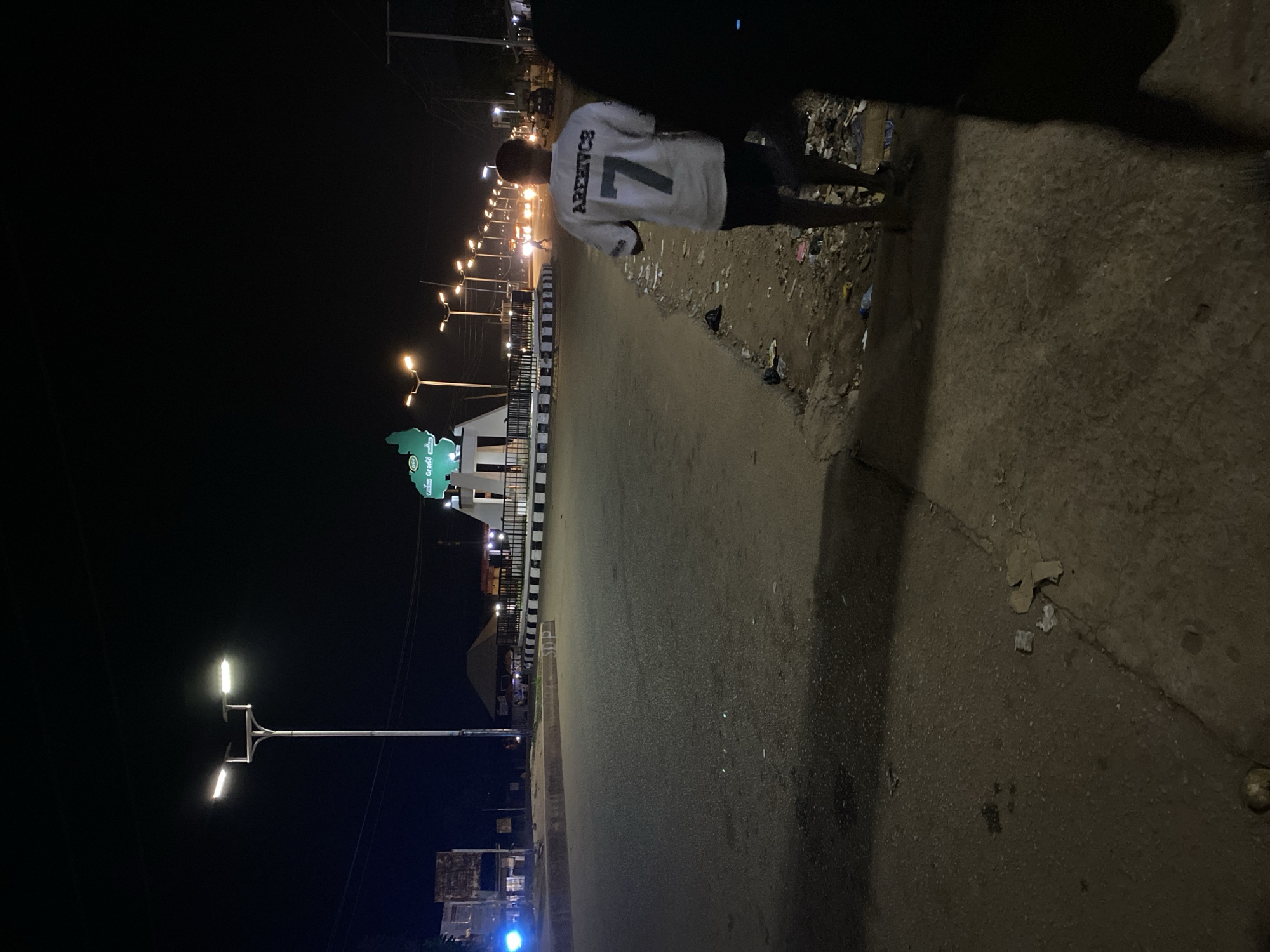 The image shows a person standing on the side of a road at night in Bukuru, Nigeria. The person is wearing a white shirt with the number "7" and the name "ARERVICS" printed on the back. The road is lit by streetlights, and there are buildings visible in the distance. The edge of the road is littered with trash. The foreground is partially obscured by a dark shadow.