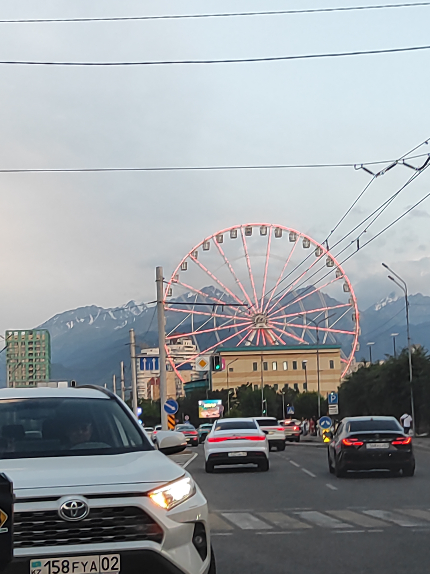 The image captures a street scene in Almaty, Kazakhstan, featuring a prominent Ferris wheel illuminated with pink lights. The Ferris wheel is situated behind a line of buildings, with the majestic snow-capped mountains serving as a backdrop. In the foreground, a white Toyota RAV4 is visible, with its headlights on, suggesting it's either dusk or an overcast day. Several other cars are on the road, indicating traffic. A pedestrian crossing is marked on the asphalt. The sky is cloudy, and overhead power lines are also present. The overall impression is an urban environment with a leisure attraction and natural scenery in the distance.