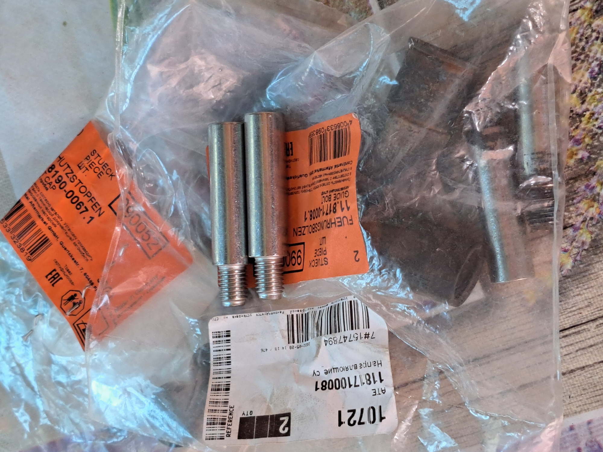 The image displays two new automotive brake caliper guide bolts, packaged within clear plastic. The bolts are silver and metallic, with threaded ends. One bolt is positioned above the other, and both are slightly angled. Attached to the packaging is a white label with black text and a barcode, indicating "REFERENCE", "QTY", and the number "2". Below this, another white label with black text reads "10721 ATE 11817100081 Направляющие су" and "7#15747894". To the right of the bolts, an orange label with black text is visible, stating "8190-0067.1 HUTZSTOPPEN", "STUECK", "PIECE", "WT.", and "CAP". Further text on the orange label includes "FUEHRUNGSBOLZEN 11.8171-0008.1 GUIDE BOLT", and a barcode. Another smaller orange label has the text "EAC". The background appears to be a wooden surface, possibly a table or floor. The overall setting suggests a retail or storage environment, and the presence of an EAC mark on the orange label indicates the product is intended for the Eurasian Economic Union market. The location of Vypasne, Ukraine, is not visually represented or suggested by any elements within the image.
