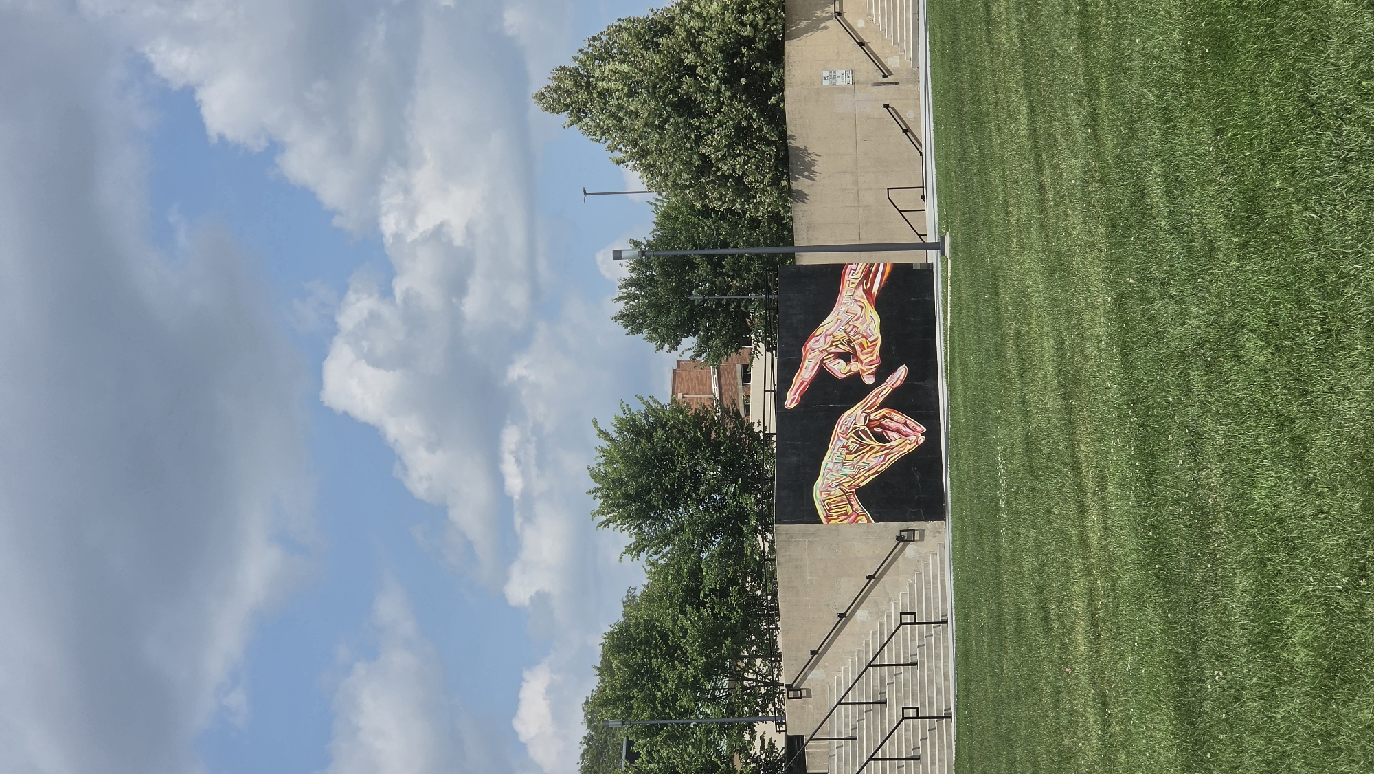 The image shows an outdoor scene on a sunny day with a blue sky and scattered white clouds. In the foreground, a lush green lawn is visible. Behind the lawn, there is a concrete wall with a large mural of two hands reaching towards each other, depicted with colorful, geometric lines. To the left and right of the mural, concrete staircases with black railings lead upwards. Several trees with dense green foliage are planted behind the wall, and a brick building can be seen in the distance. Streetlights are also present in the scene. The mural's style suggests it's a contemporary piece of public art, possibly located in a park or public plaza within the city of Elgin, United States of America.