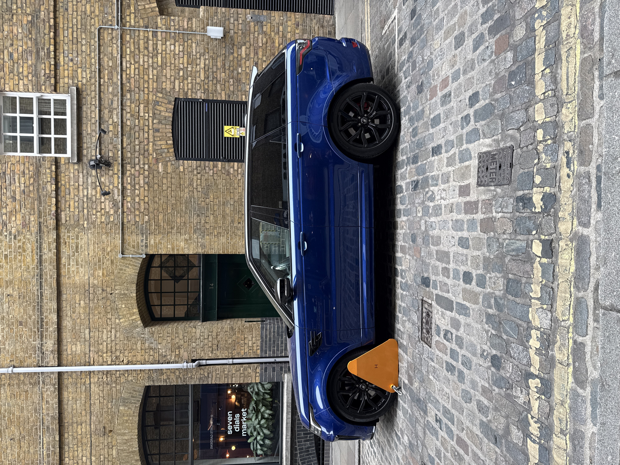 A blue Range Rover has been clamped in a narrow street in London, United Kingdom. The car is parked on a cobblestone road marked with a faded yellow line. The surrounding architecture consists of a brick building with several arched windows and a green door with the number 14. A sign for "Seven Dials Market" is visible through a ground-level window. Overhead, a window with white framing is visible. A black metal fixture with a yellow warning sign hangs on the exterior wall.