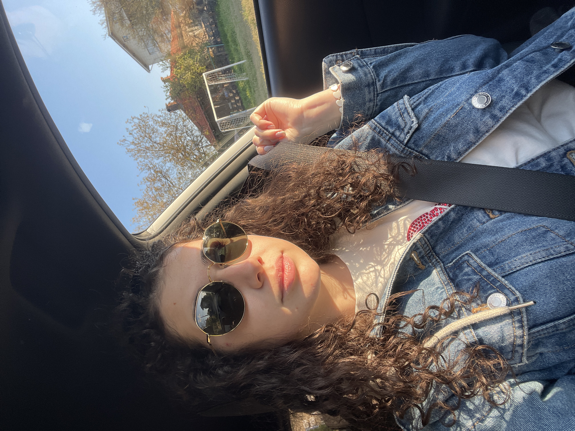 The image captures a young woman, likely a passenger, inside a car on a bright, sunny day in Sofia, Bulgaria. She has prominent curly brown hair and wears aviator-style sunglasses. Her face is illuminated by strong sunlight, creating highlights and shadows, and her lips appear to have a glossy finish. She is secured by a black seatbelt.
She wears a blue denim jacket over a white top, which has a red and white sequined pattern visible near her chest. The silver buttons on her denim jacket clearly display the word "TRUST." Her left hand, adorned with a white heart-shaped beaded bracelet and light pink manicured nails, rests near the window or sunroof opening.
Through the car's windows, a clear blue sky is visible. The exterior scene includes lush green grass, deciduous trees, distant houses—one with a white facade and a red-tiled roof—and a white soccer goal in what appears to be a field or large yard. The strong sunlight and clear sky suggest a pleasant outdoor day, possibly morning or late afternoon given the angle of the light. The car's interior appears dark, contrasting with the bright outdoor scene.